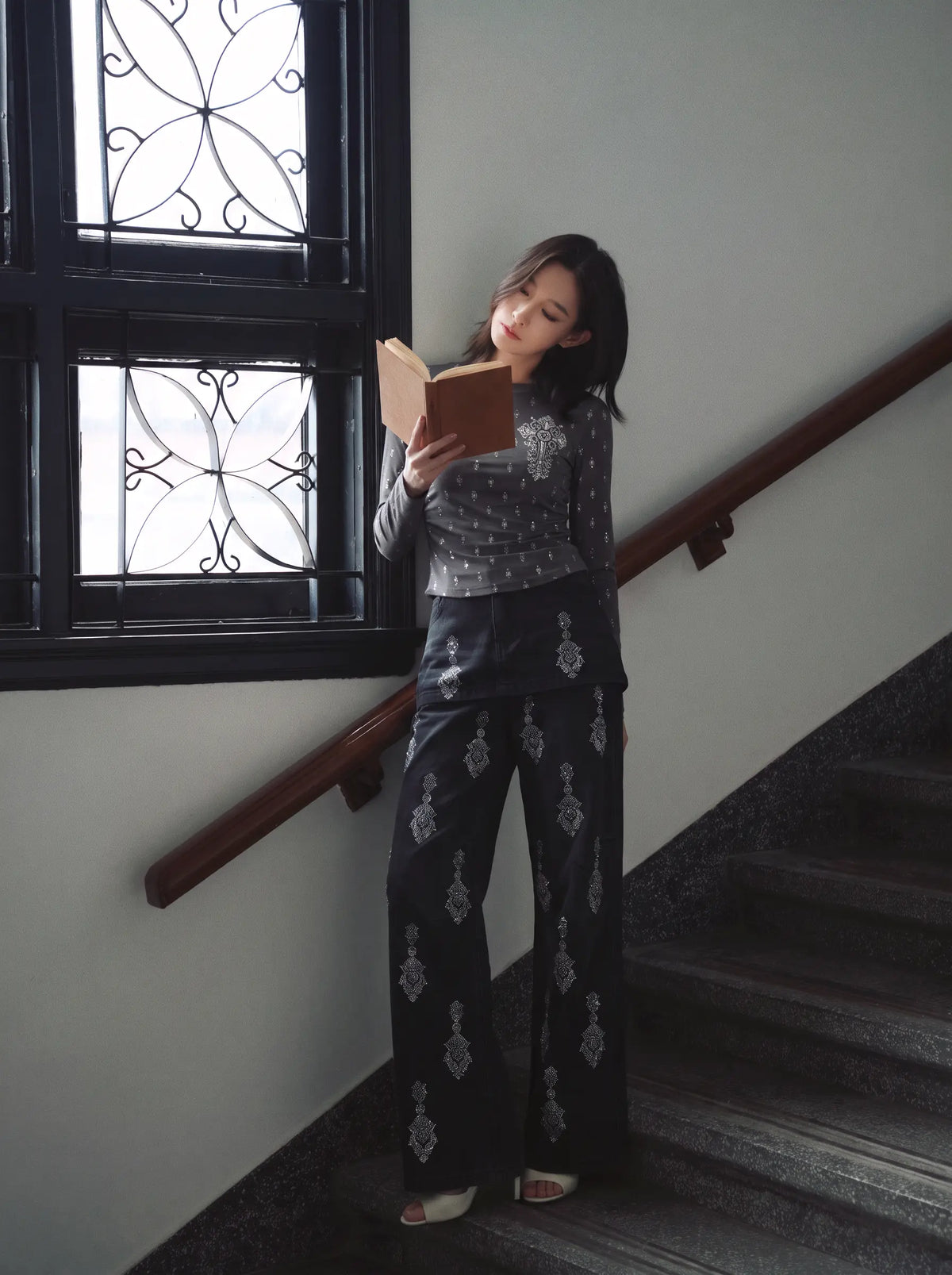 A woman stands on an indoor staircase near a window, holding and reading a book. She is wearing a matching two-piece outfit with a dark gray patterned long-sleeve top and dark pants featuring silver-toned ornamental motifs. TCH Rhinestone Cross Graphic Fitted Long Sleeve Top