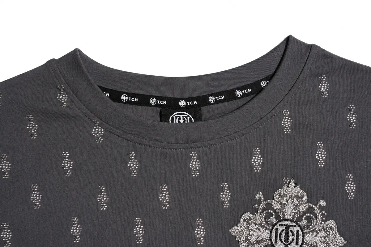 Close-up of a dark gray crewneck t-shirt with a repeating small silver rhinestone pattern and a large silver rhinestone crest on the lower right chest. The inner collar shows the brand name 'TCM' printed repeatedly on a black label. TCH Rhinestone Cross Graphic Fitted Long Sleeve Top
