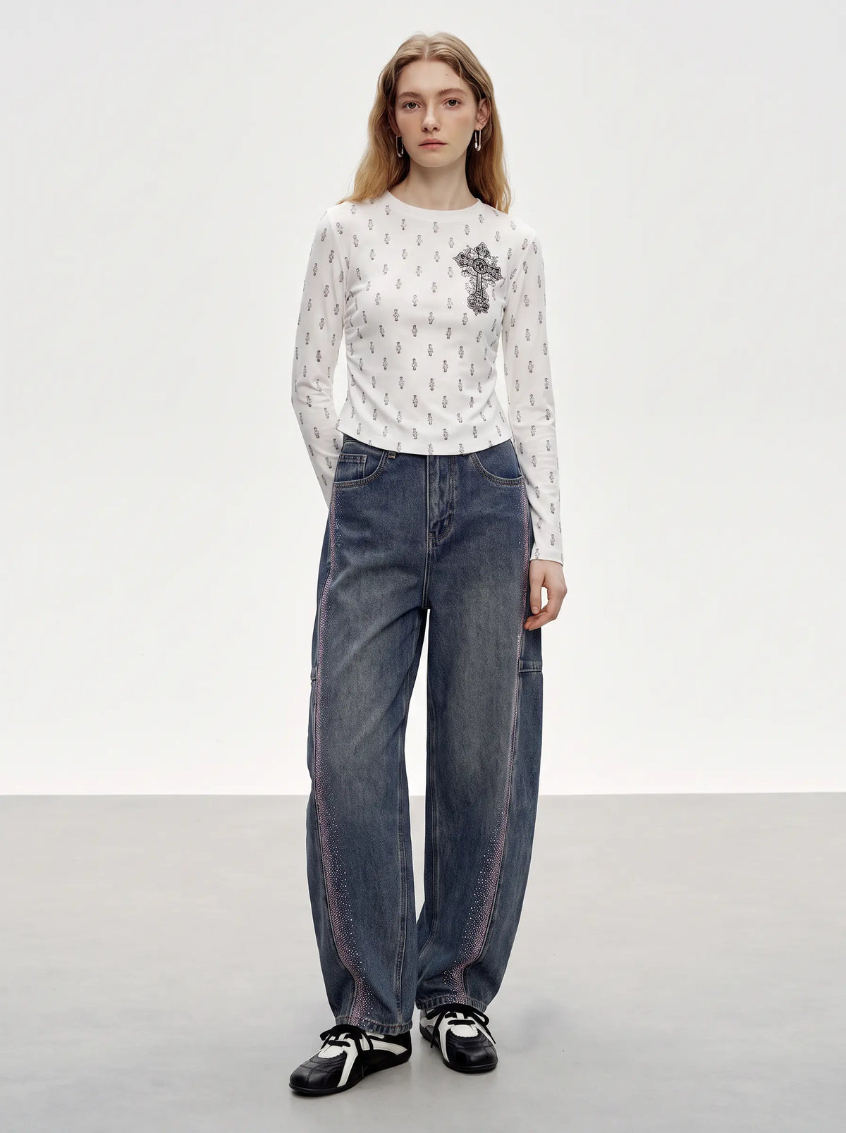 A model stands in a studio wearing a white long-sleeve knit top with a small black cross motif and dark blue wide-leg jeans. TCH Rhinestone Cross Graphic Fitted Long Sleeve Top