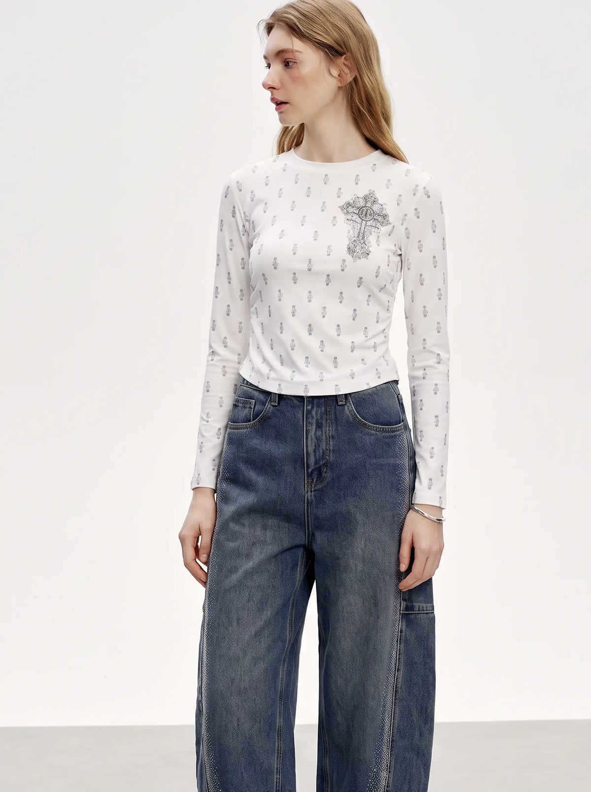 A woman modeling a white long-sleeve crewneck top with a small gray monogram pattern and a gray embroidered cross motif on the left chest, paired with high-waisted blue jeans against a plain white studio background. TCH Rhinestone Cross Graphic Fitted Long Sleeve Top