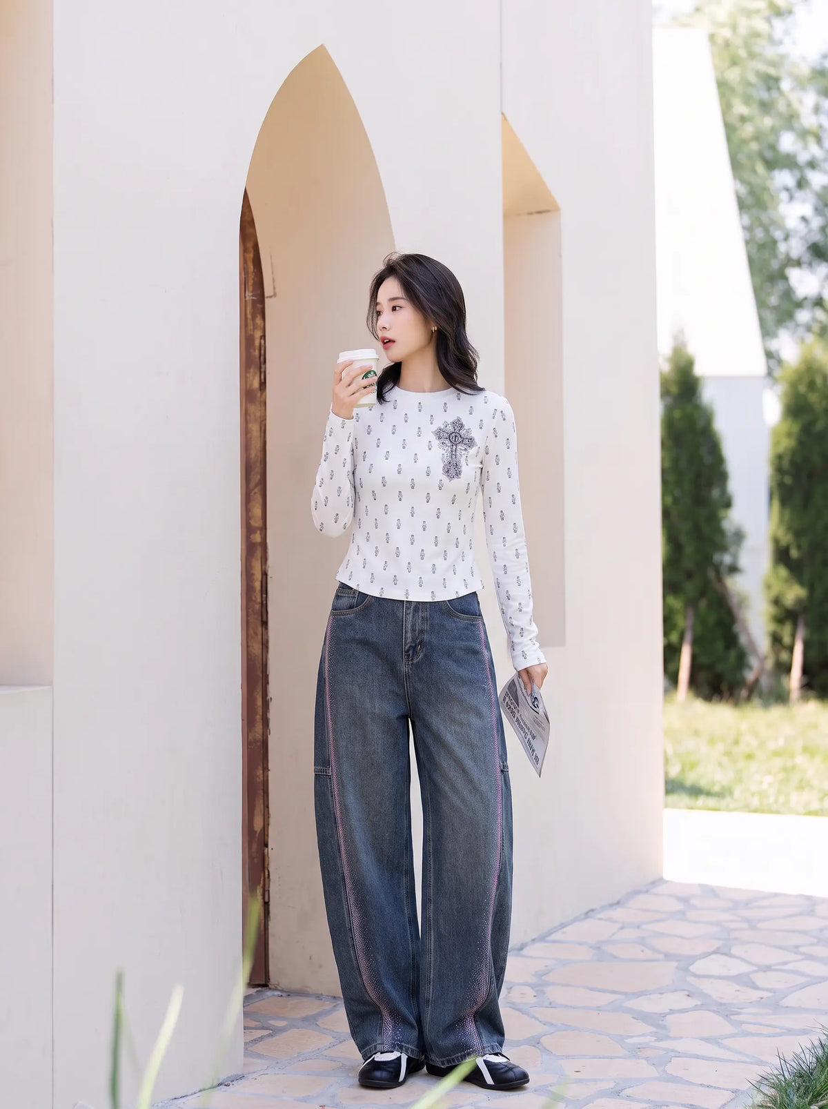 A woman stands outdoors in front of a beige arched wall, wearing a white long-sleeve top with a small gray geometric pattern and dark wide-leg jeans. She is holding a white coffee cup. TCH Rhinestone Cross Graphic Fitted Long Sleeve Top