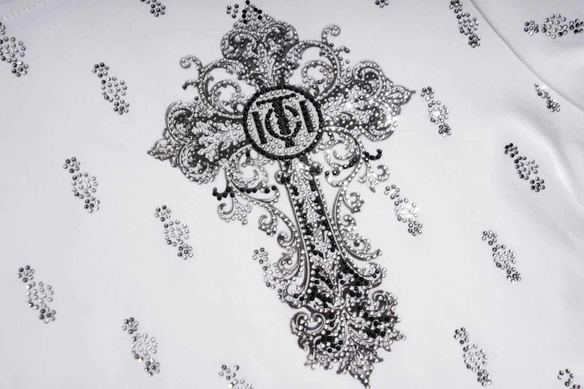 A close-up of a white garment featuring a large ornate black embroidered cross with a circular monogram logo in the center, surrounded by smaller matching embroidered motifs. TCH Rhinestone Cross Graphic Fitted Long Sleeve Top