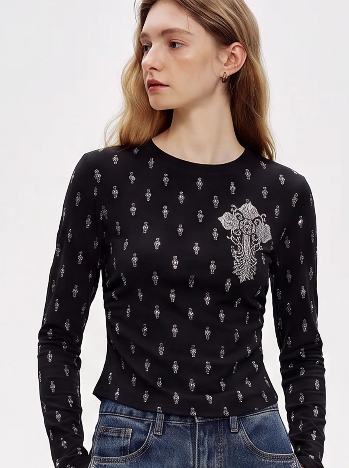 A woman wearing a fitted black long-sleeve top with a small repeating silver graphic print and a larger silver floral emblem on the left chest, paired with blue jeans against a light gray studio background. TCH Rhinestone Cross Graphic Fitted Long Sleeve Top