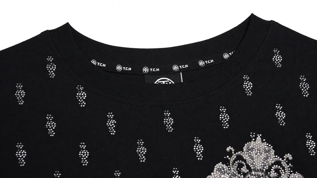 Close-up of a black crewneck top with silver rhinestone embellishments, featuring a repeating vertical dot pattern and a large ornate crown motif on the lower right chest. The inner neck label shows the brand name TCM. TCH Rhinestone Cross Graphic Fitted Long Sleeve Top