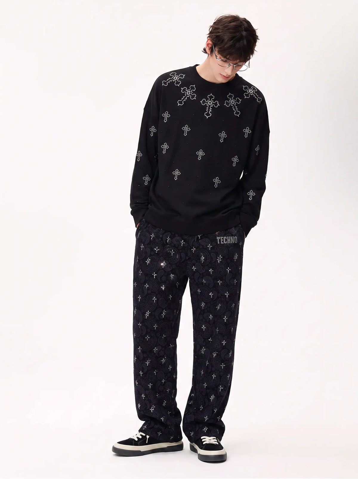 A model wearing a black long-sleeve crewneck sweatshirt and matching black sweatpants, both featuring a repeating white cross pattern. The sweatshirt has a small white graphic on the chest, and the pants have a small white text graphic on the thigh. TCH Rhinestone Cross Pattern Black Long Sleeve T-Shirt