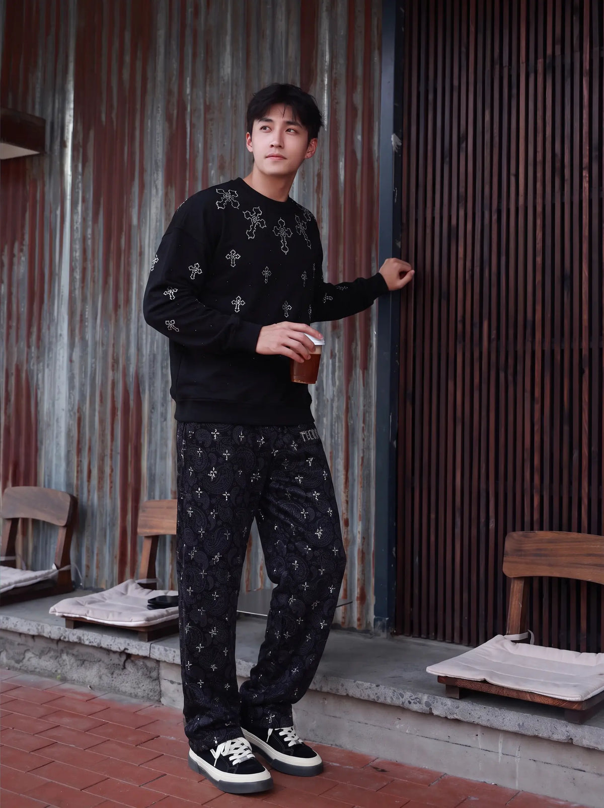 A man wearing a black long-sleeve crewneck sweater and matching black patterned pants, standing in front of a rustic corrugated metal and wood slat wall while holding a brown paper cup. TCH Rhinestone Cross Pattern Black Long Sleeve T-Shirt