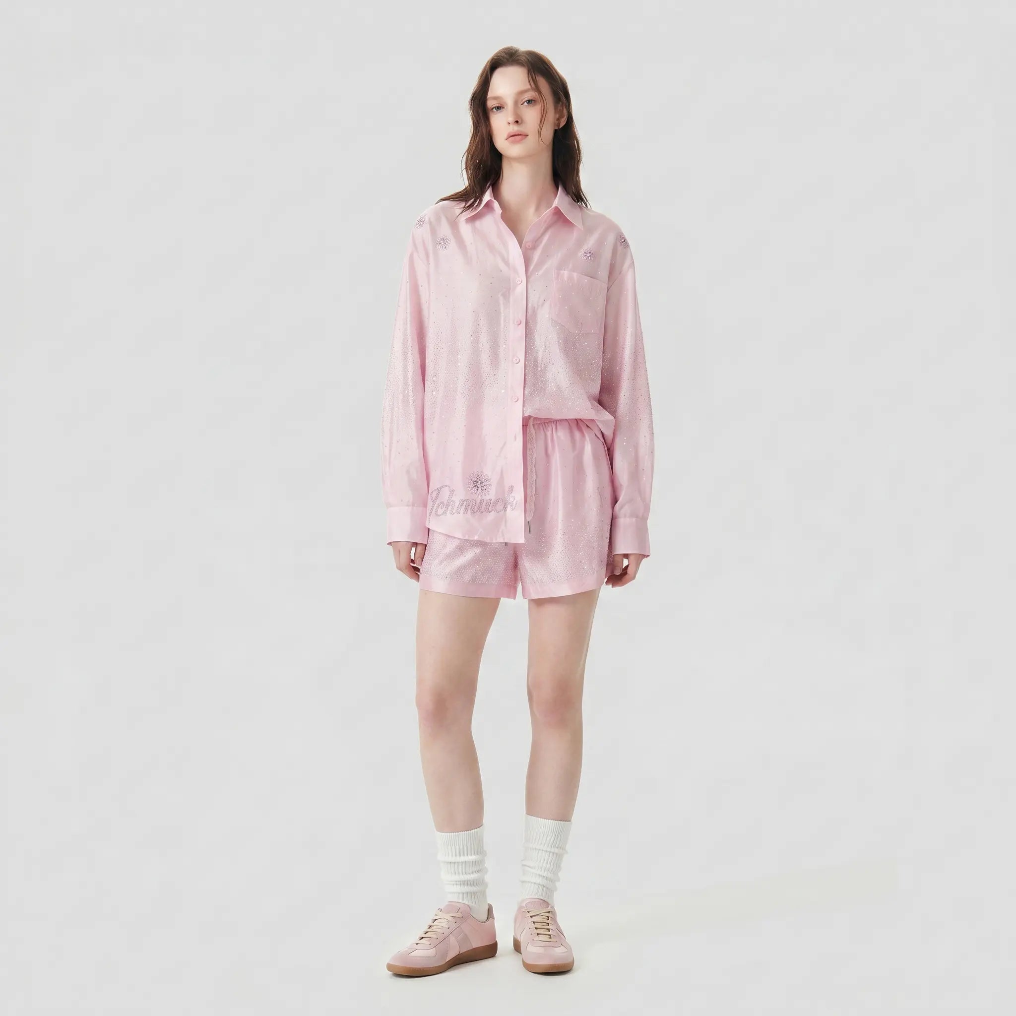 A model stands against a light gray studio background wearing a light pink matching lounge set consisting of a long-sleeve button-up shirt and shorts, paired with white socks and pink slip-on shoes. TCH Rhinestone Crystal Embellished Pink Shirt & Shorts Set