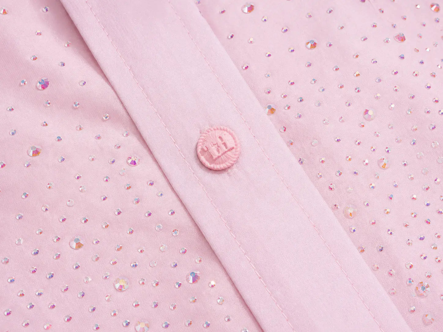 Close-up of a light pink garment with a diagonal seam, a matching pink button, and scattered iridescent rhinestones on a pink fabric background. TCH Rhinestone Crystal Embellished Pink Shirt & Shorts Set