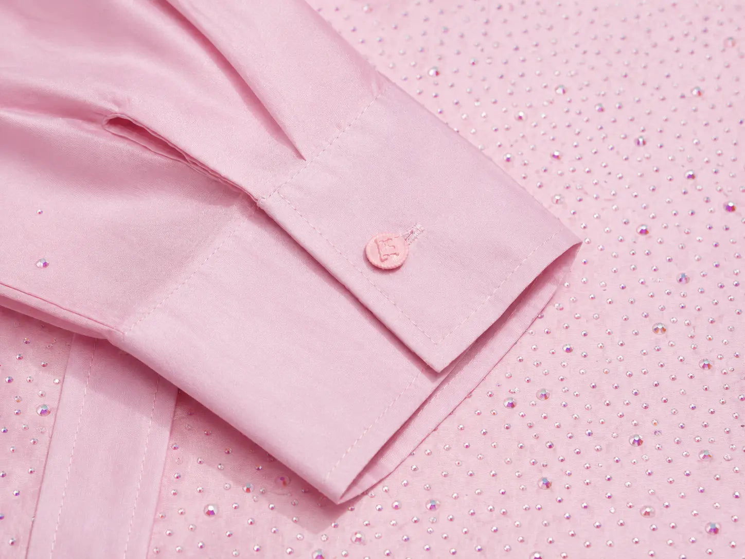 A close-up of a light pink dress shirt sleeve with a buttoned cuff, laid on top of a matching pink fabric surface covered with small rhinestones. TCH Rhinestone Crystal Embellished Pink Shirt & Shorts Set