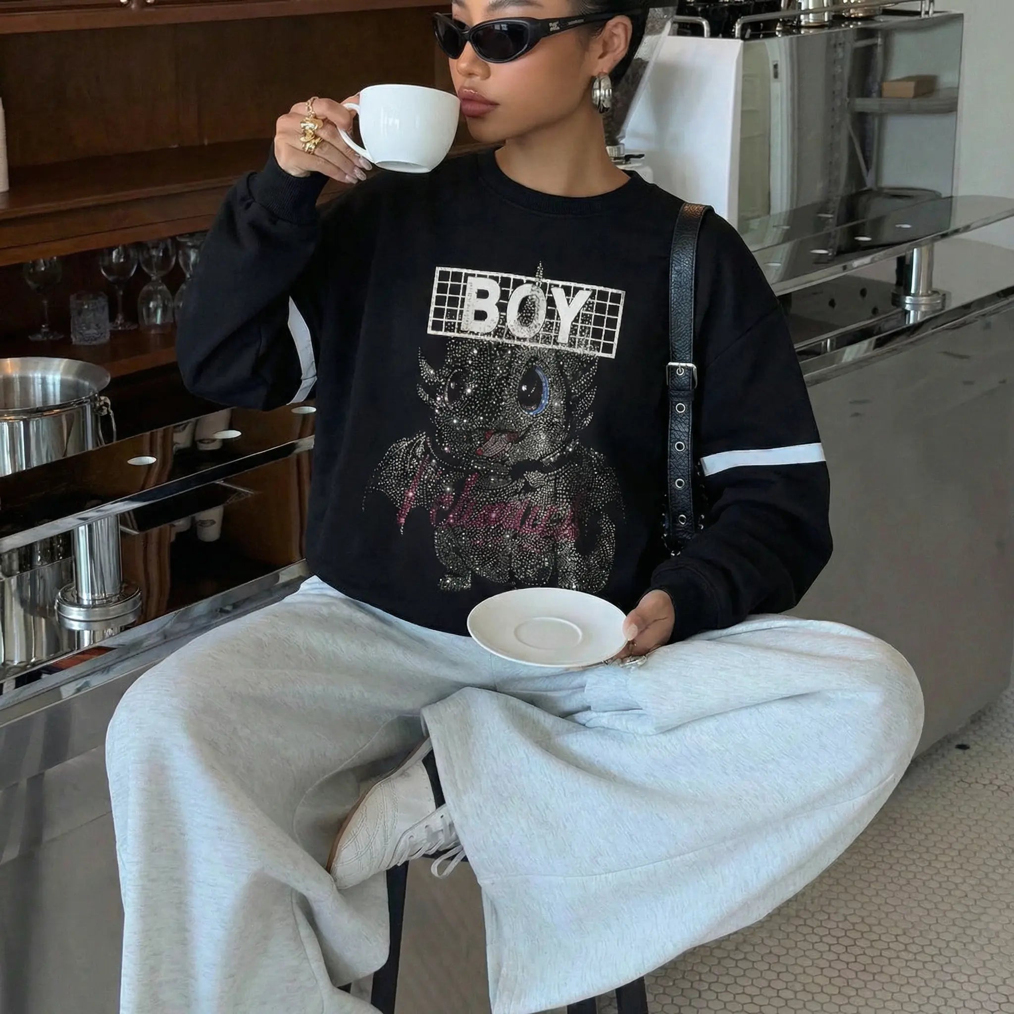 Person sitting in a cafe holding a cup and saucer, wearing a black sweatshirt with 'BOY' graphic design. TCH Rhinestone Cute Dragon Sweatshirt