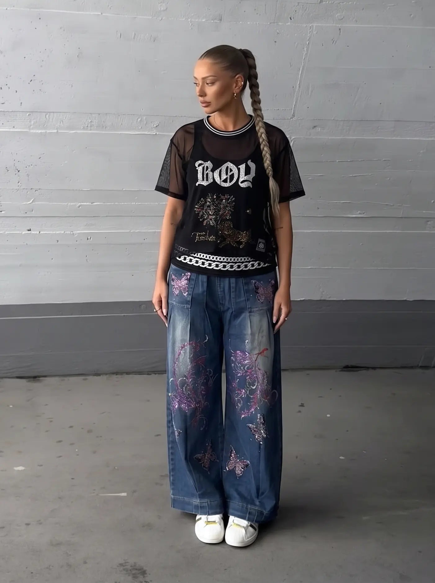 A model stands against a gray concrete wall wearing a black graphic t-shirt with 'BOY' in gothic letters, wide-leg blue jeans with purple butterfly embroidery, and white sneakers. TCH Rhinestone Deer Graphic Mesh Sleeve Black Streetwear T-Shirt