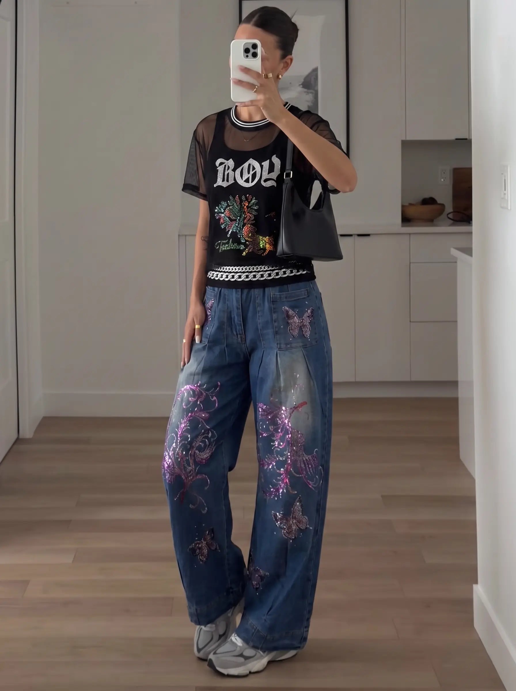A person takes a mirror selfie in a room with light wood floors and white cabinets, wearing a sheer black graphic t-shirt, wide-leg blue jeans with purple butterfly embellishments, and gray sneakers. TCH Rhinestone Deer Graphic Mesh Sleeve Black Streetwear T-Shirt