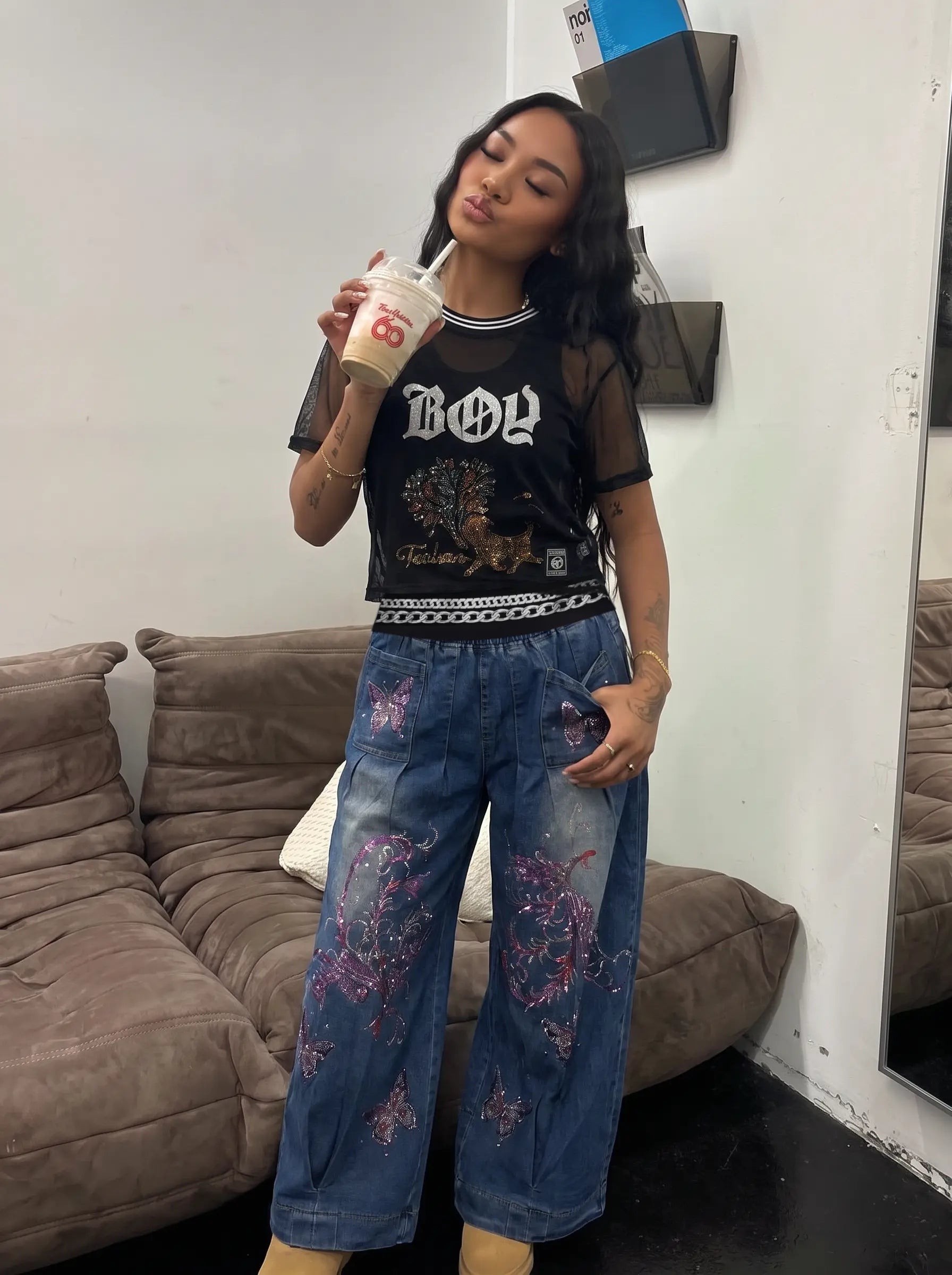 A woman posing in a black sheer-sleeve graphic t-shirt and blue denim jeans with pink floral embroidery, holding a drink in a living room setting. TCH Rhinestone Deer Graphic Mesh Sleeve Black Streetwear T-Shirt