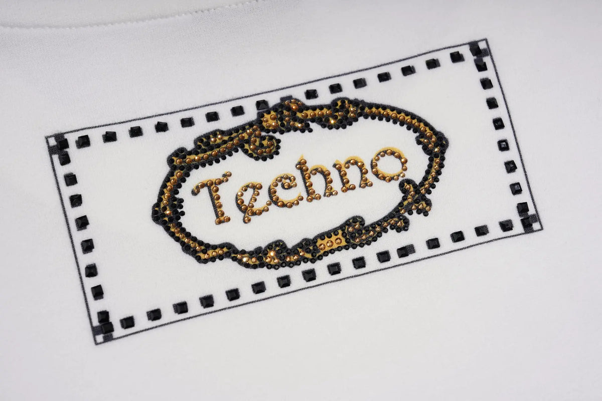 A close-up of a white garment featuring an embroidered rectangular patch with a black dotted border and the word 'Techno' in gold lettering inside an ornate gold frame. TCH Rhinestone Denim Corset Graphic Long Sleeve T-Shirt