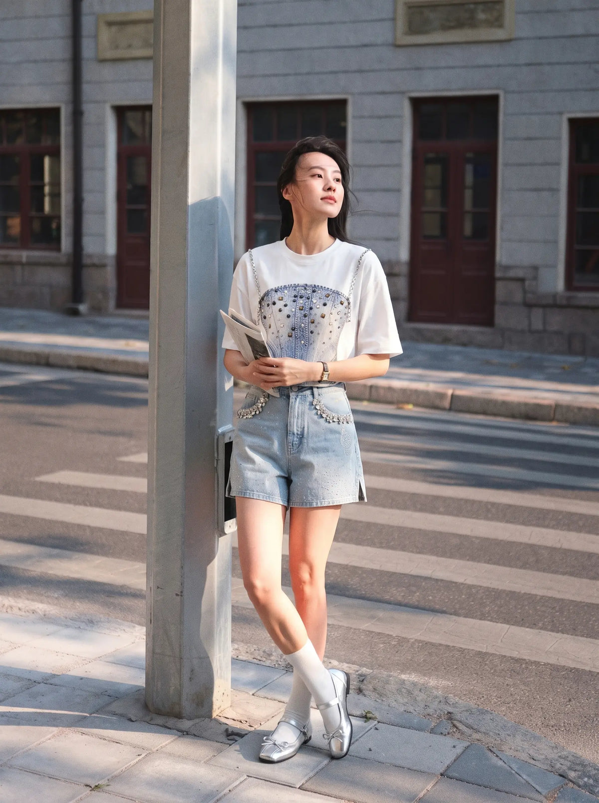 A woman stands on a city sidewalk next to a pole, wearing a white graphic T-shirt with a rhinestone-embellished chest design, light-wash denim shorts with silver studs, white socks, and white loafers. The background shows a street and a gray stone building. TCH Rhinestone Denim Corset Graphic Long Sleeve T-Shirt