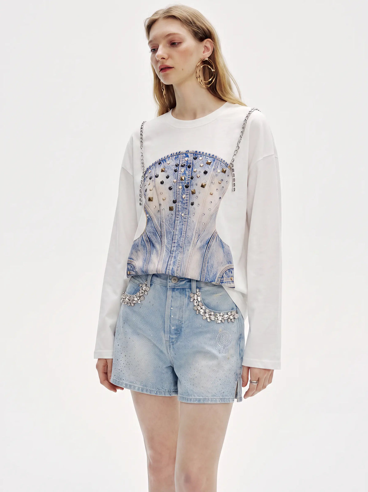 A model wearing a white long-sleeve crewneck top with a blue denim corset graphic print, paired with light-wash denim shorts featuring silver rhinestone embellishments, against a plain white studio background. TCH Rhinestone Denim Corset Graphic Long Sleeve T-Shirt