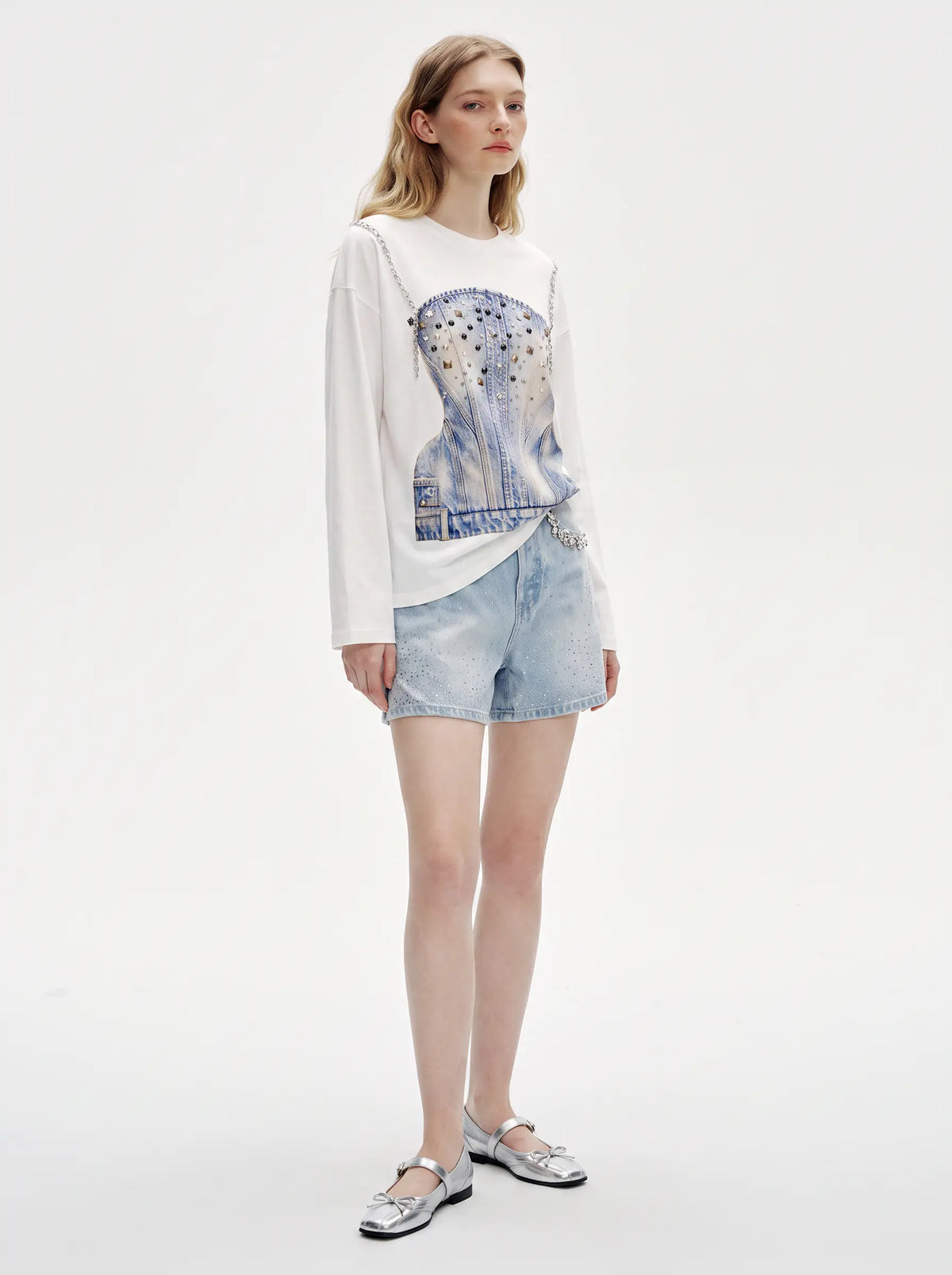 A model stands against a plain white studio background wearing a white long-sleeve top with a blue denim corset graphic, light-wash denim shorts, and silver flat shoes. TCH Rhinestone Denim Corset Graphic Long Sleeve T-Shirt