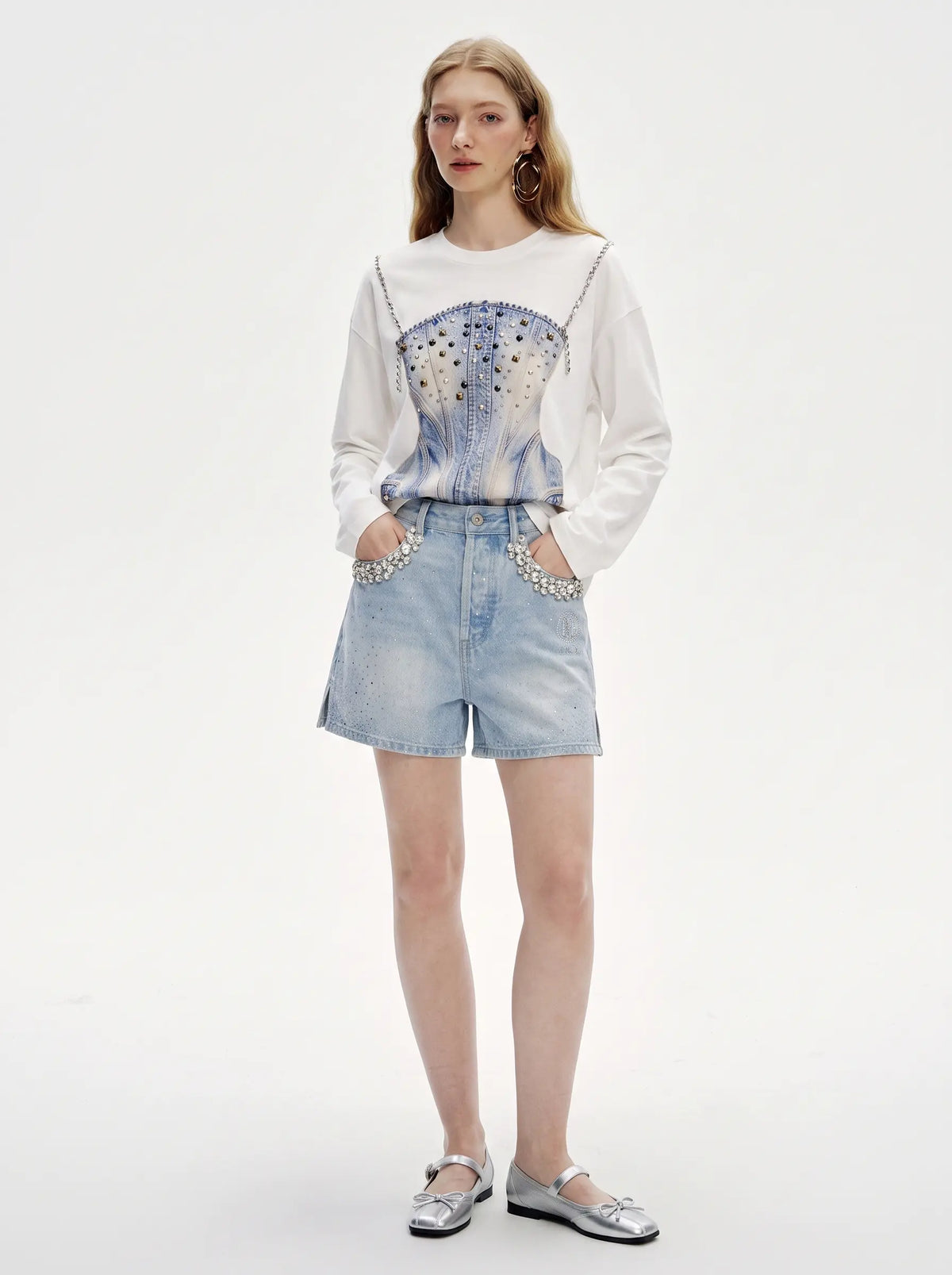 A model stands against a plain white studio background wearing a white long-sleeve top with a blue and silver graphic print, light-wash denim shorts with silver rhinestone trim, and silver flats. TCH Rhinestone Denim Corset Graphic Long Sleeve T-Shirt
