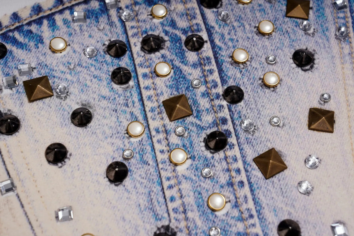 Close-up of a light blue and off-white denim jacket heavily embellished with black and gold studs, faux pearls, and clear rhinestones. TCH Rhinestone Denim Corset Graphic Long Sleeve T-Shirt