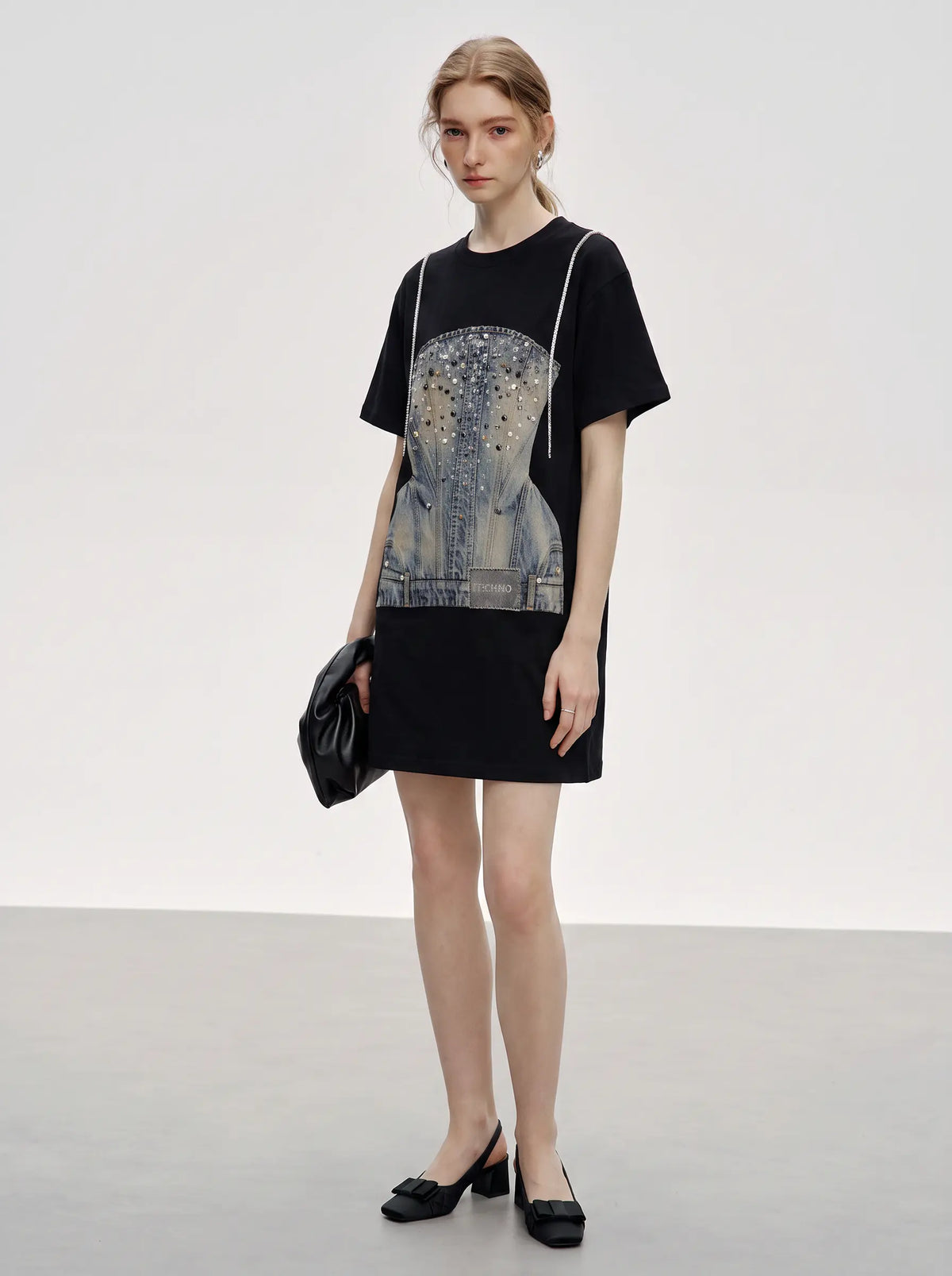 A model stands in a studio wearing a black oversized T-shirt dress with a large blue-gray graphic print on the front, black pointed flats, and holding a black clutch bag. TCH Rhinestone Denim Corset Graphic Oversized T-Shirt