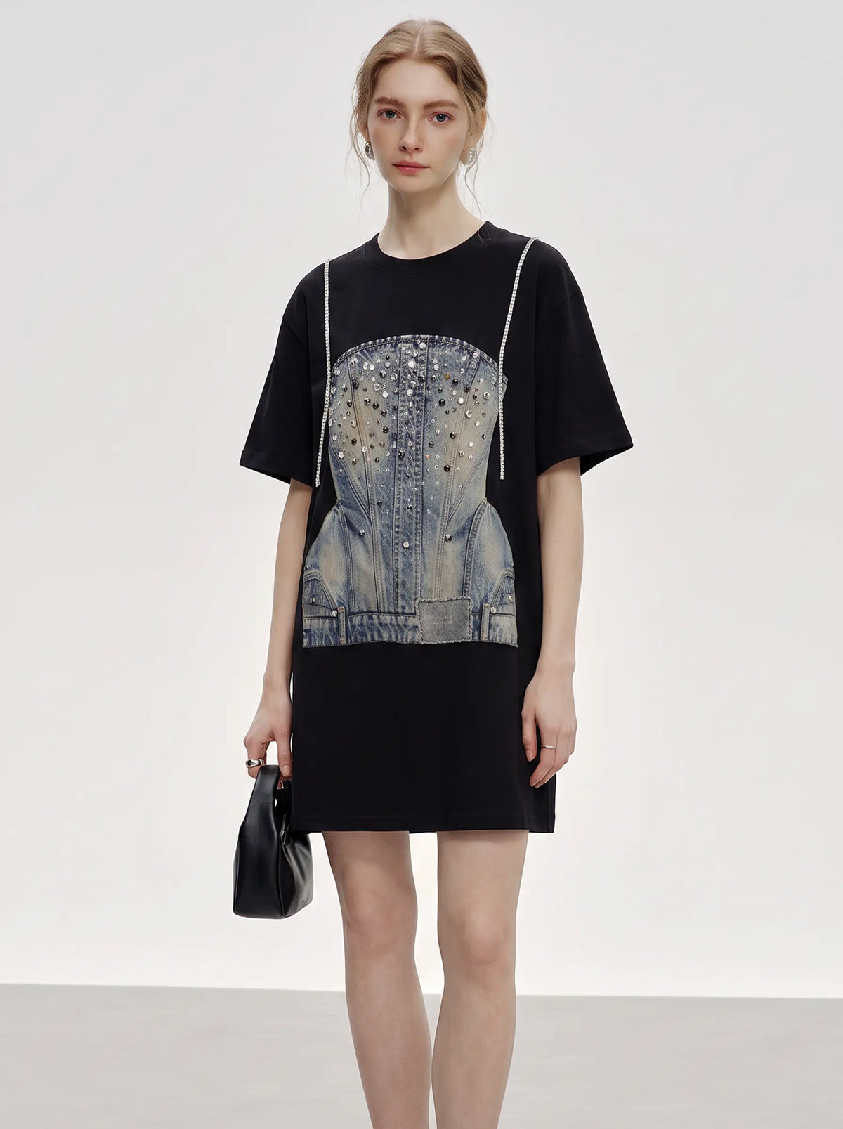 A model wearing a black oversized t-shirt dress with a denim corset graphic print on the front, holding a small black handbag against a plain light gray studio background. TCH Rhinestone Denim Corset Graphic Oversized T-Shirt