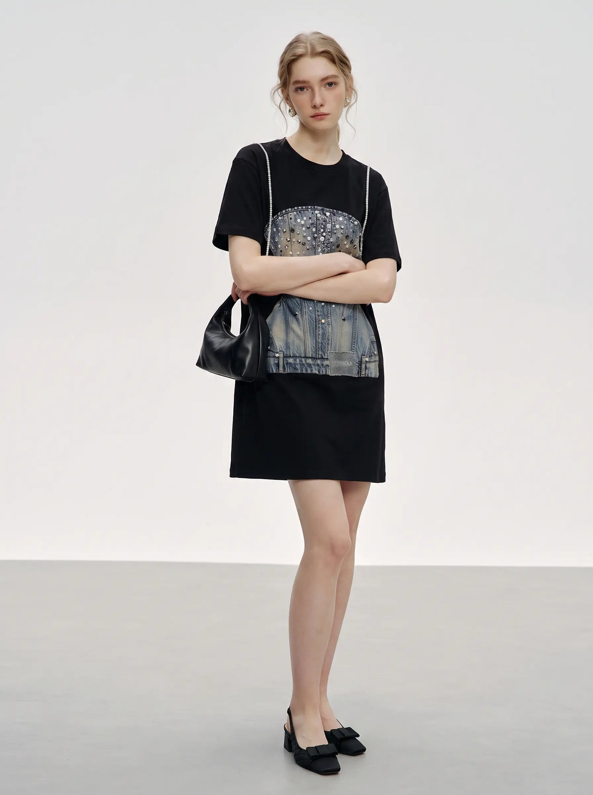 A model stands in a studio wearing a black mini dress with a large denim and sequined graphic panel on the front, paired with black pointed flats and a black handbag. TCH Rhinestone Denim Corset Graphic Oversized T-Shirt