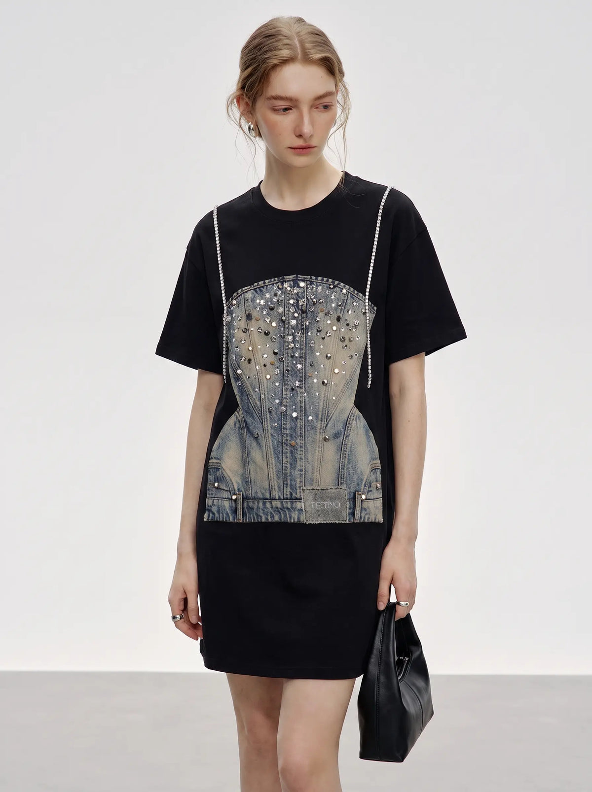 A model wearing a black short-sleeve mini dress with a large denim corset graphic embellished with rhinestones on the front, holding a black leather handbag against a light gray studio background. TCH Rhinestone Denim Corset Graphic Oversized T-Shirt