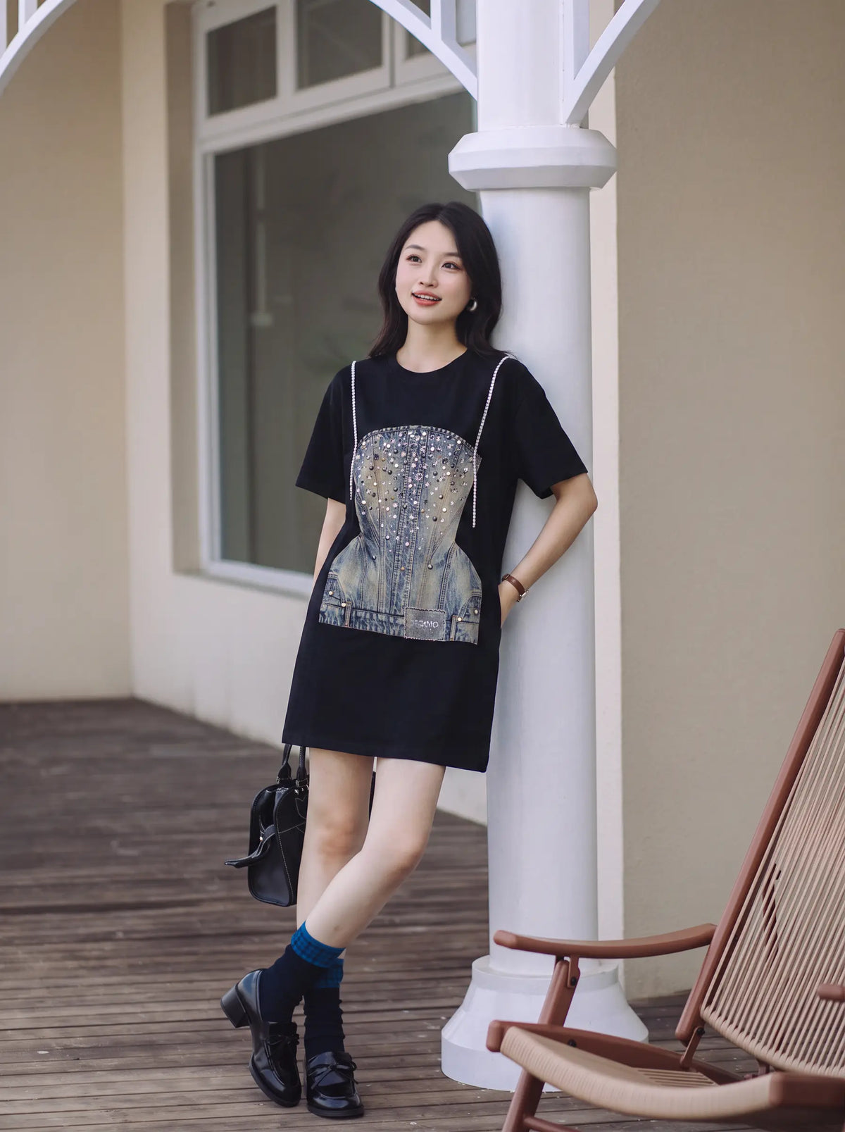 A woman stands on a wooden porch wearing a black short-sleeve mini dress with a large blue and gold graphic print on the front. She is leaning against a white column, holding a black handbag, with a wooden chair visible in the foreground. TCH Rhinestone Denim Corset Graphic Oversized T-Shirt