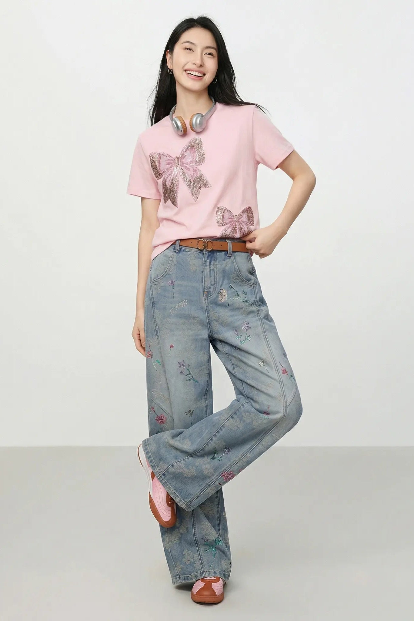 A woman stands against a plain light gray studio background wearing a light pink graphic T-shirt with butterfly motifs, wide-leg light-wash blue jeans with small floral embroidery, a brown belt, and brown sandals. She has headphones around her neck. TCH Rhinestone Double Bow Cotton T-Shirt