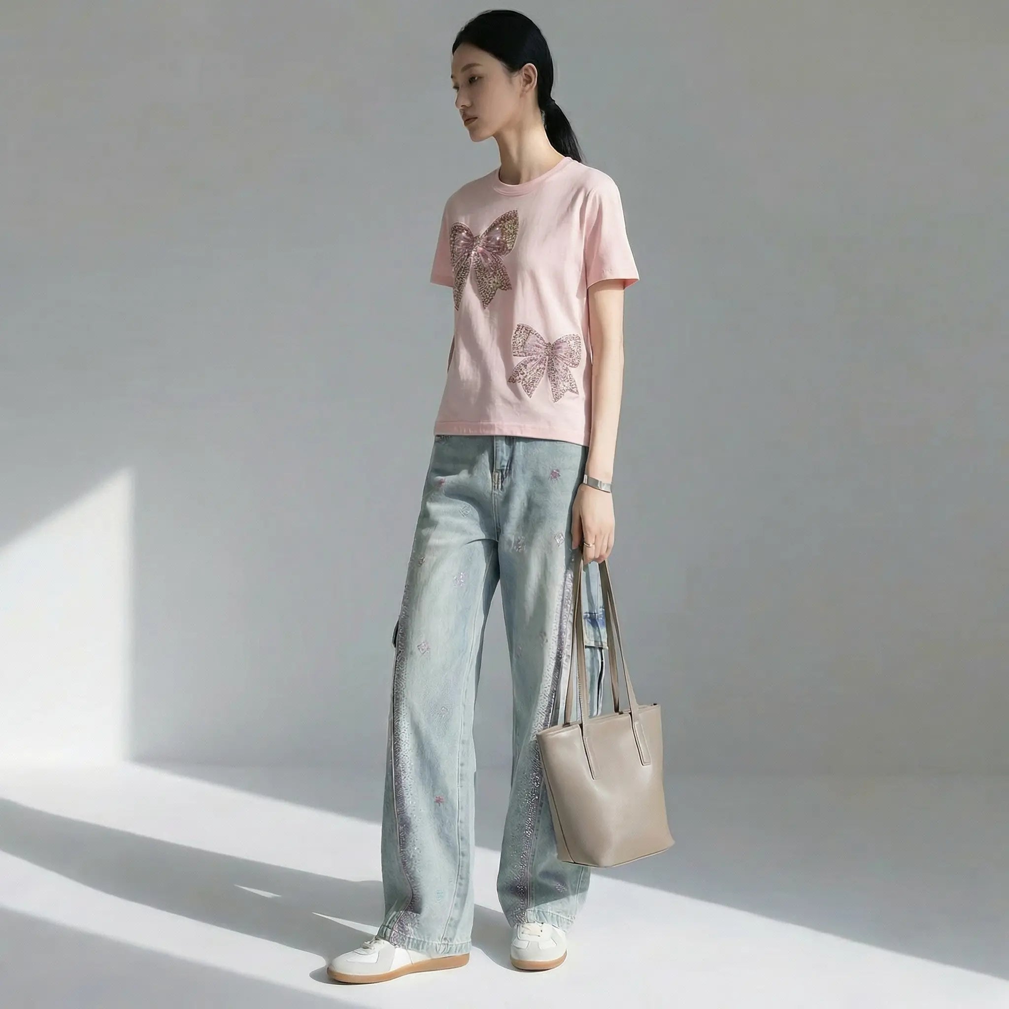 A woman stands in a studio wearing a light pink short-sleeve T-shirt with two large sequined butterfly motifs, light-wash wide-leg jeans, white sneakers, and holding a taupe tote bag. TCH Rhinestone Double Bow Cotton T-Shirt