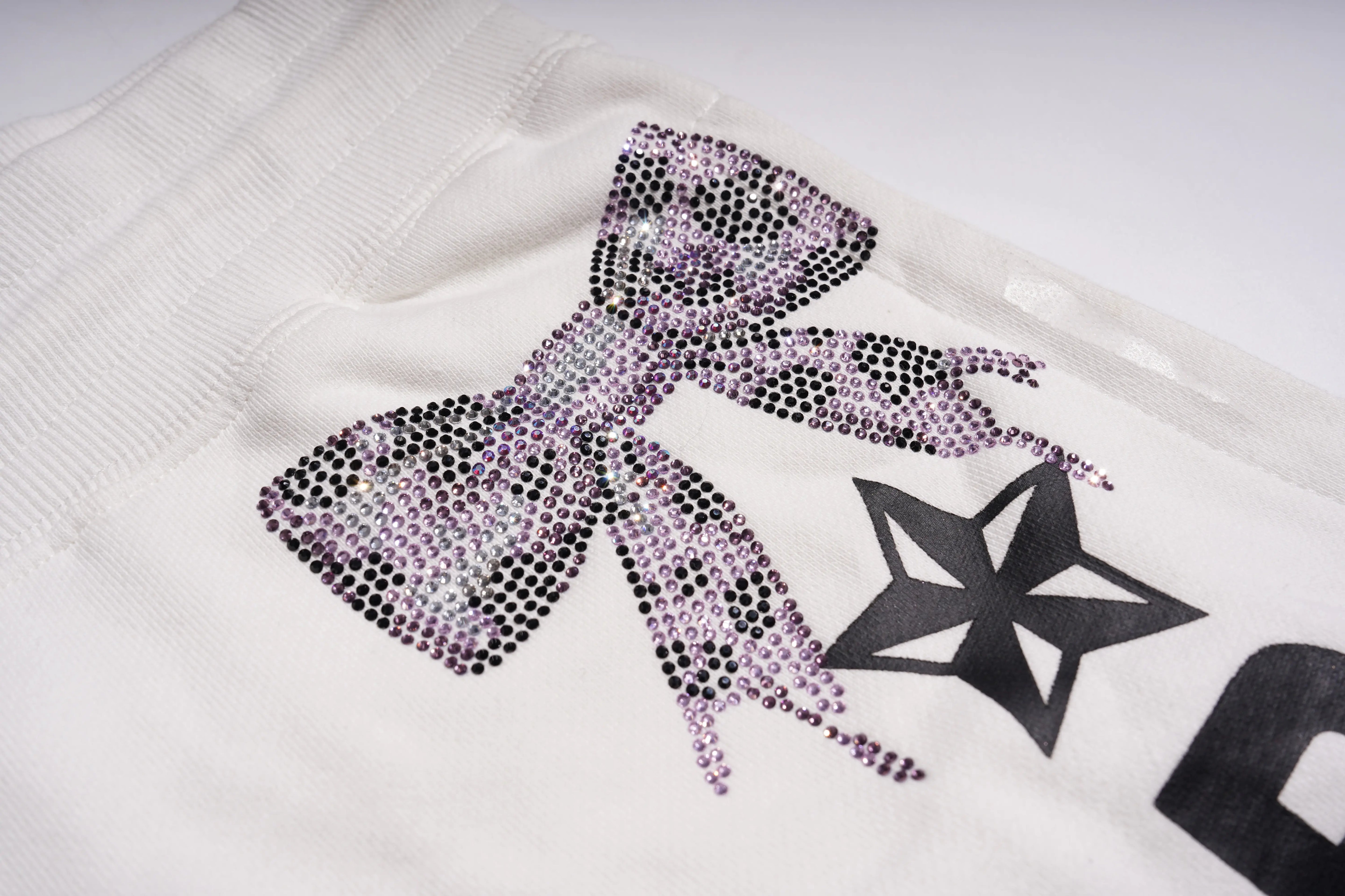 Close-up of a white sweater with a purple rhinestone bow and black star design. TCH Rhinestone Elephant Bow White Casual Shorts Luxury Streetwear Brand Official Showcase