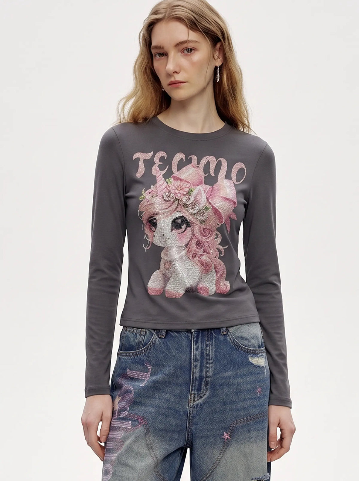A woman modeling a dark gray long-sleeve t-shirt with a pink and white cartoon unicorn graphic and the word 'TECMO' across the chest, paired with distressed blue jeans against a plain white background. TCH Rhinestone Fantasy Unicorn Graphic Long Sleeve T-Shirt 