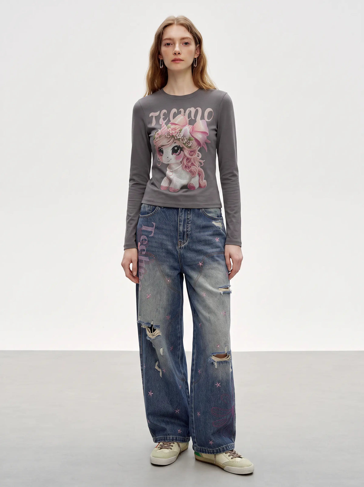 A model stands against a light gray studio background wearing a dark gray long-sleeve graphic t-shirt with a pink cartoon unicorn print and the word 'ecmo', paired with distressed blue jeans and beige sneakers. TCH Rhinestone Fantasy Unicorn Graphic Long Sleeve T-Shirt 