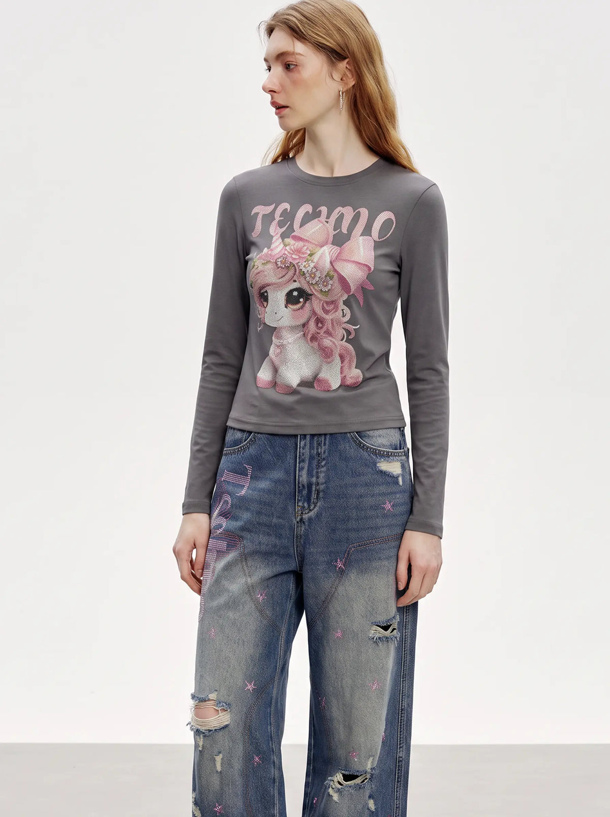 A woman modeling a dark gray long-sleeve t-shirt with a pink cartoon unicorn graphic and the word 'TERMO' printed across the chest, paired with distressed blue jeans. TCH Rhinestone Fantasy Unicorn Graphic Long Sleeve T-Shirt