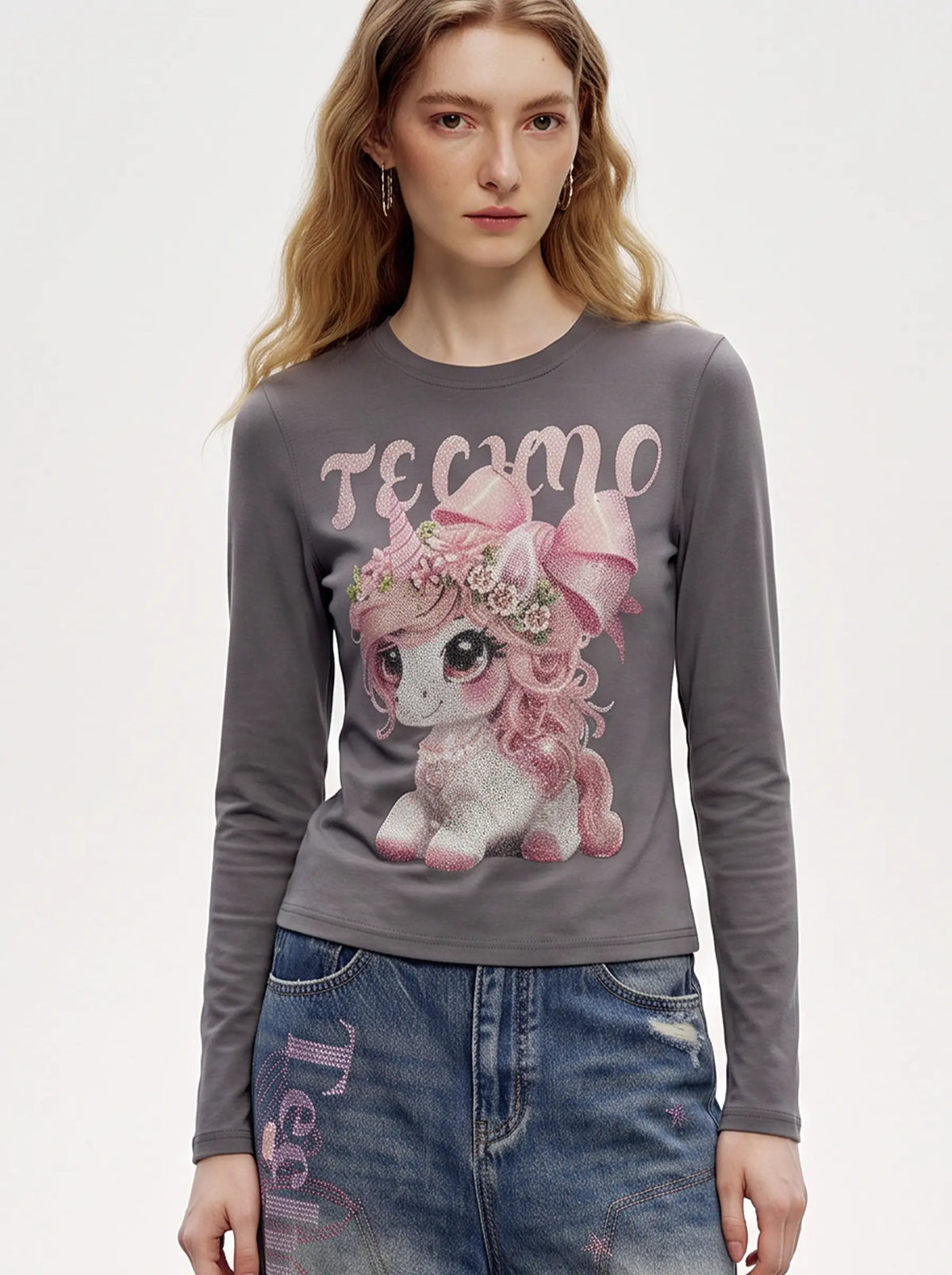 A model wearing a light blue denim jacket with various embroidered patches, including a teddy bear on the left chest and cherries on the sleeve, styled over a white t-shirt and dark pants. TCH Rhinestone Fantasy Unicorn Graphic Long Sleeve T-Shirt
