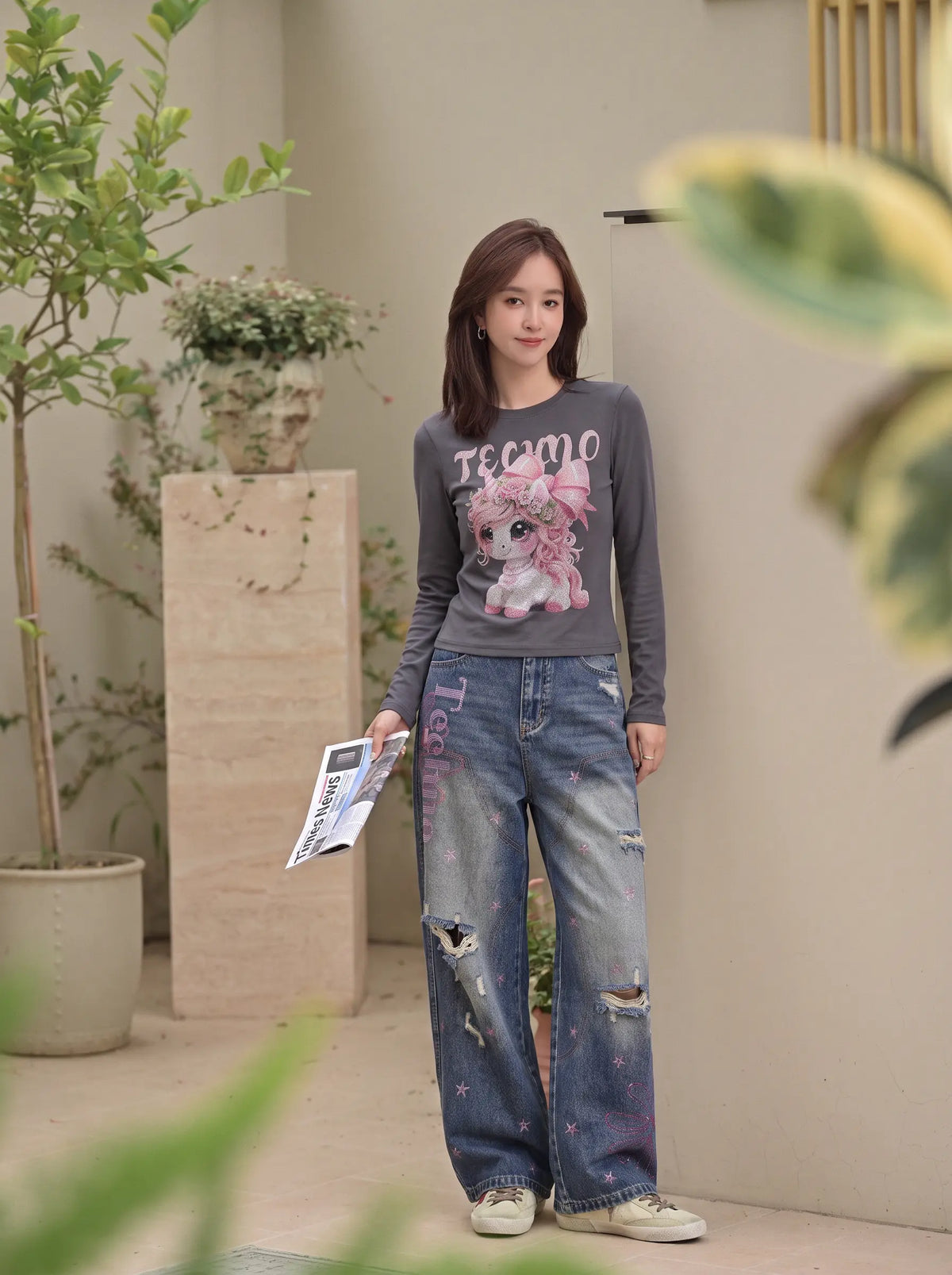 A woman stands in a modern indoor courtyard wearing a dark gray long-sleeve graphic t-shirt with a pink cartoon character and the word 'TEACHO', paired with distressed blue jeans and white sneakers. TCH Rhinestone Fantasy Unicorn Graphic Long Sleeve T-Shirt