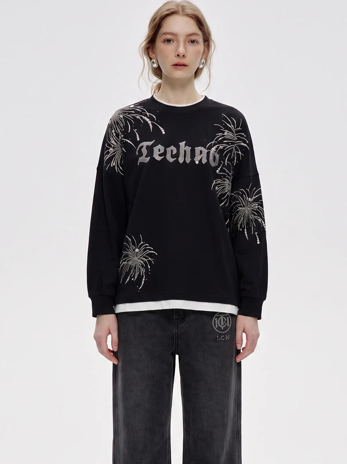 A model wearing a black crewneck sweatshirt with 'Techno' written in a gothic font across the chest, decorated with white and silver fireworks patterns on the sleeves and body. She is also wearing dark gray jeans against a plain light gray studio background. TCH Rhinestone Firework Graphic Black Sweatshirt