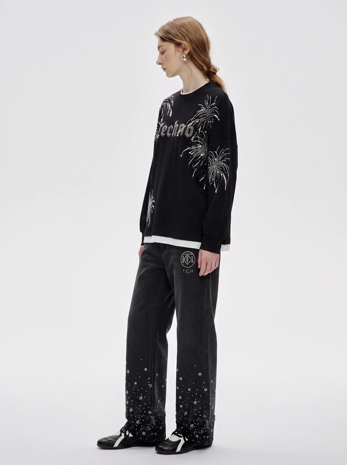 A model stands in profile wearing a black long-sleeve sweatshirt with white graphic prints and text, paired with black pants featuring white speckled details near the hem, against a plain light gray studio background. TCH Rhinestone Firework Graphic Black Sweatshirt