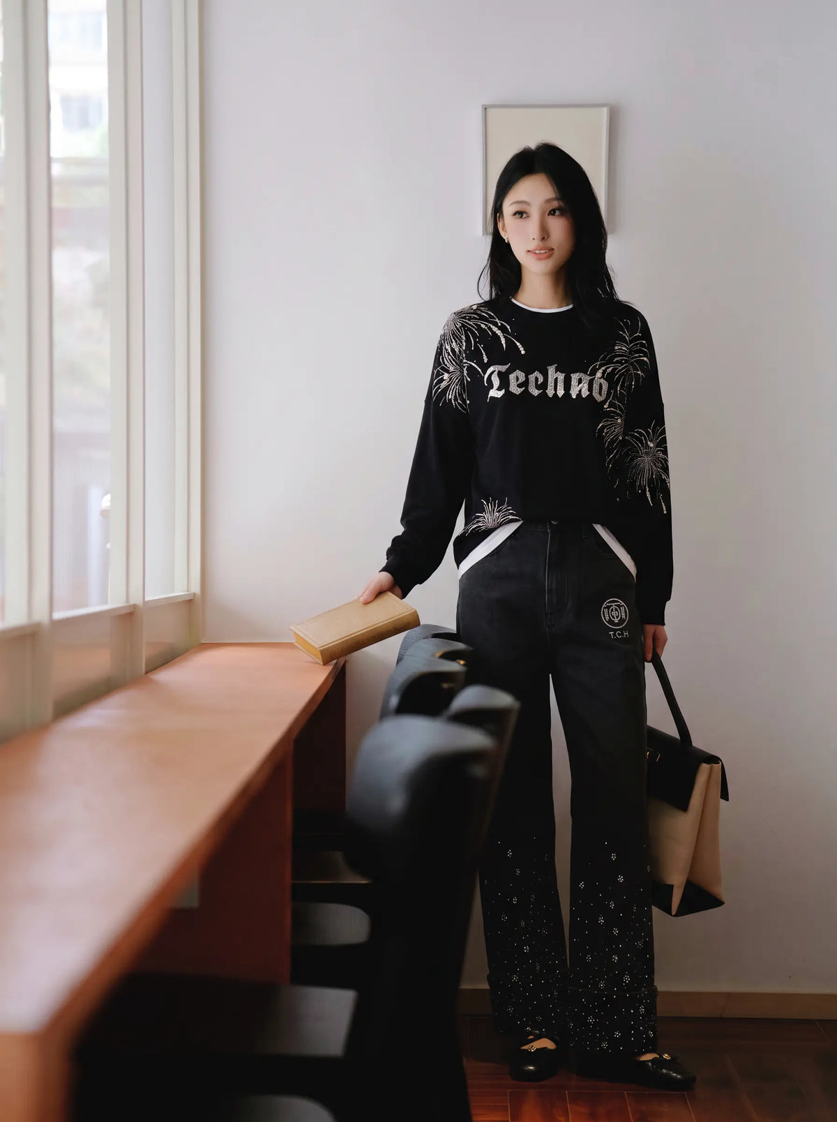 A woman stands indoors near a window and wooden table, wearing a black long-sleeve sweatshirt with white graphic text and leafy print, black pants with white side panels and speckled lower legs, and holding a tan tote bag. TCH Rhinestone Firework Graphic Black Sweatshirt