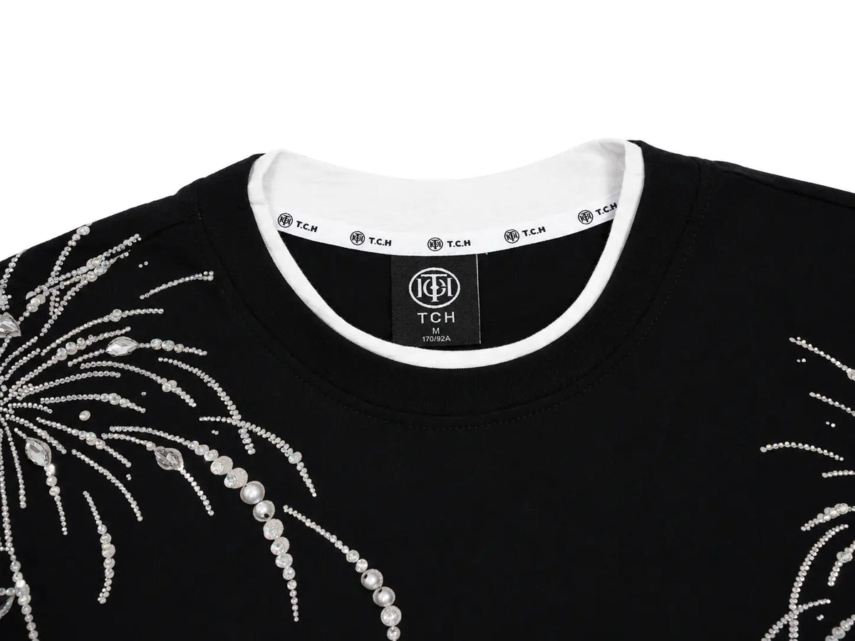 Close-up of a black crewneck sweatshirt with white pearl and rhinestone embroidery on the chest, featuring a white ribbed collar and a visible TCH brand label. TCH Rhinestone Firework Graphic Black Sweatshirt