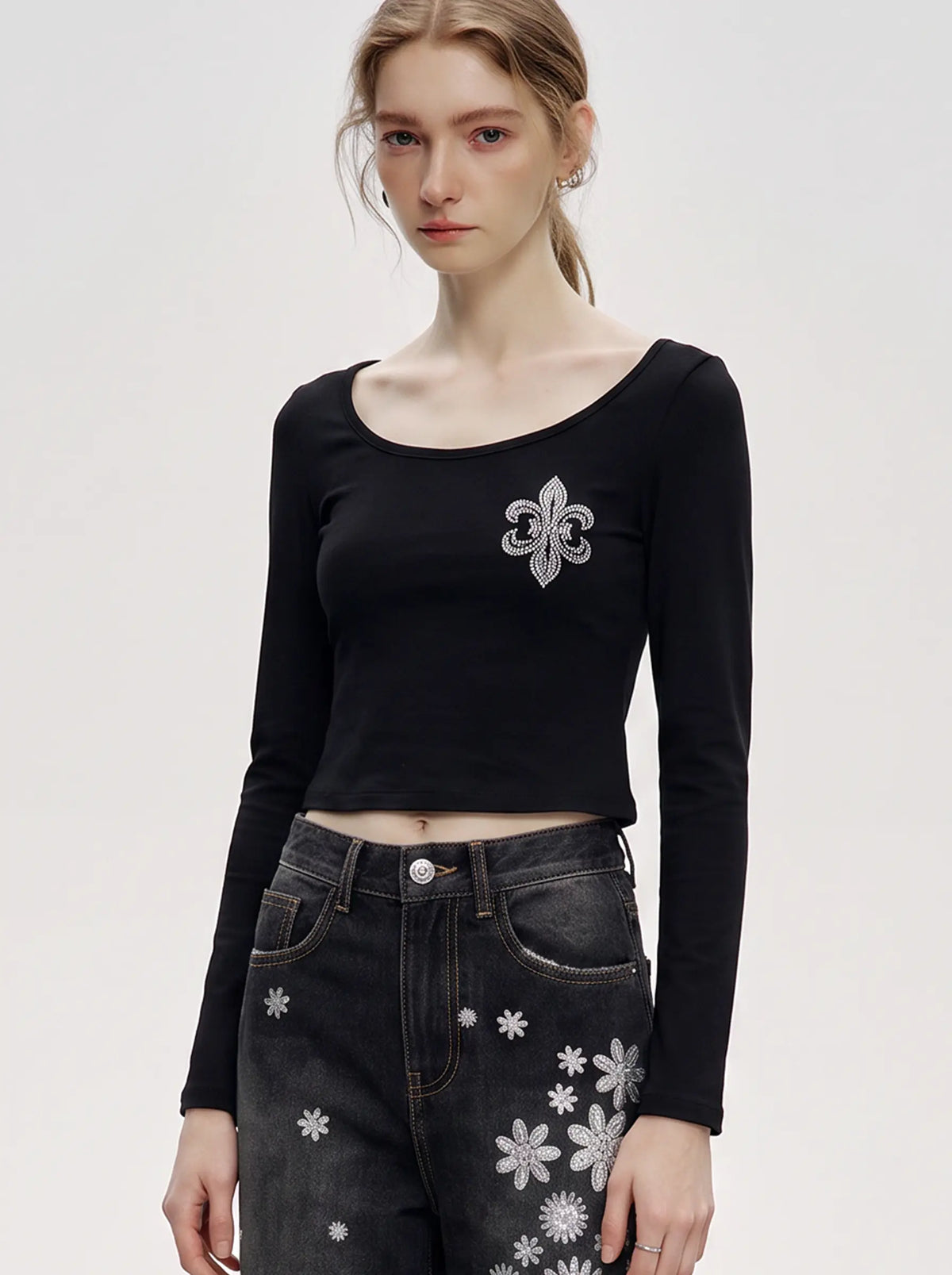 A model wearing a light blue denim jacket with various embroidered patches, including a teddy bear on the left chest and cherries on the sleeve, styled over a white t-shirt and dark pants. TCH Rhinestone Fleur-de-Lis Fitted Long Sleeve Top