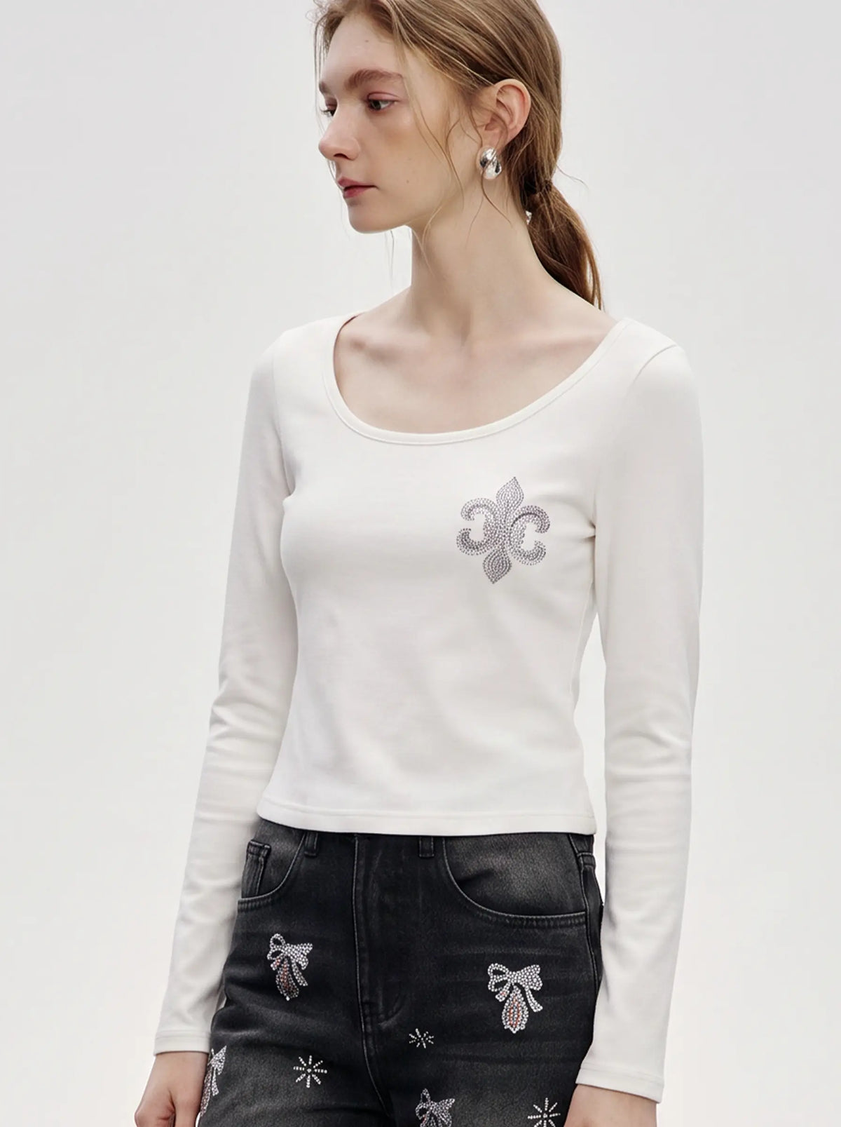 A woman wearing a fitted white long-sleeve top with a scoop neckline and a silver fleur-de-lis embellishment on the left chest, paired with dark jeans featuring bow and floral embroidery. TCH Rhinestone Fleur-de-Lis Fitted Long Sleeve Top