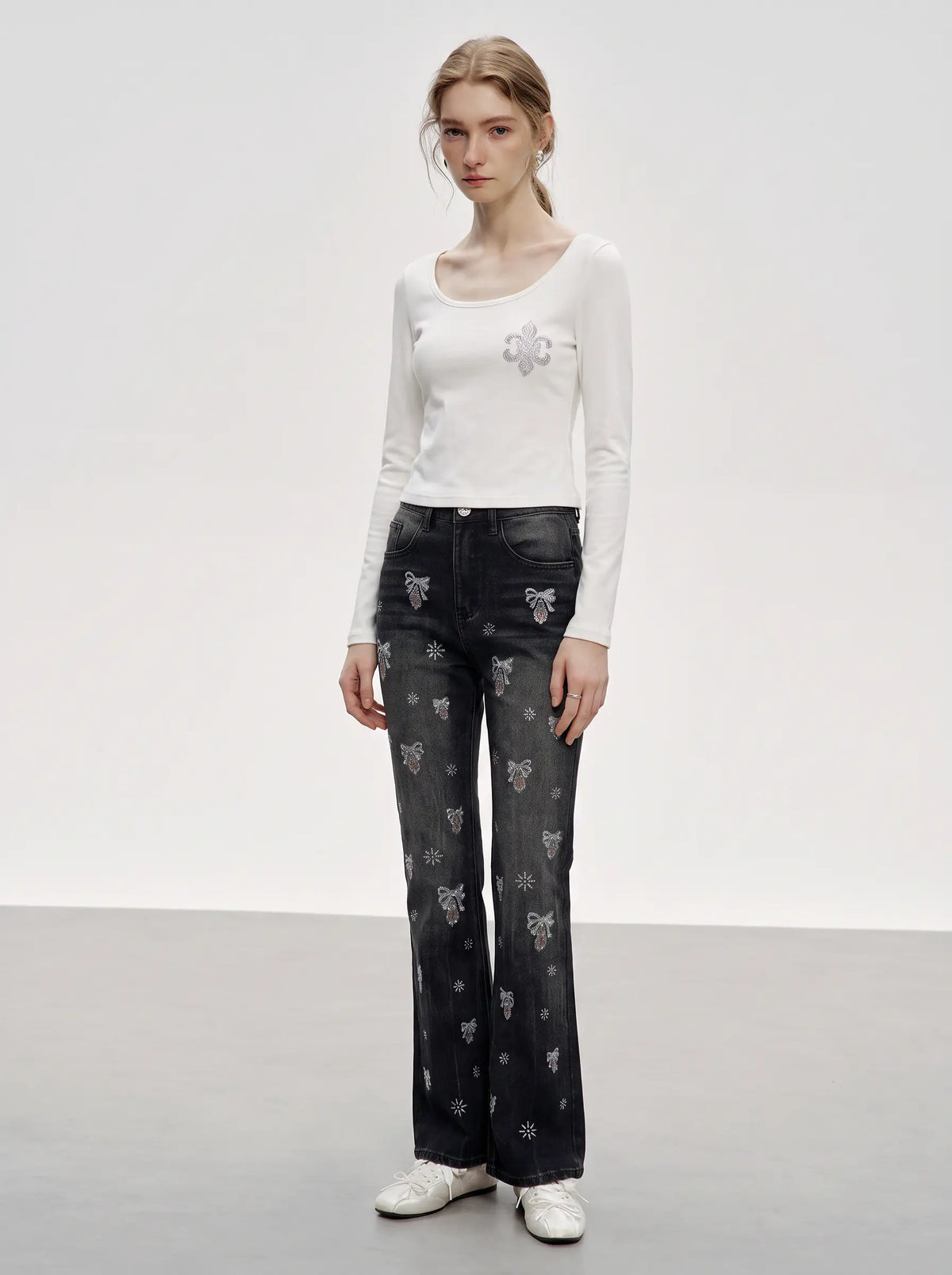 A model stands in a studio wearing a fitted white long-sleeve top with a small floral applique on the chest, dark gray wide-leg jeans with a repeating bow pattern, and white sneakers. TCH Rhinestone Fleur-de-Lis Fitted Long Sleeve Top