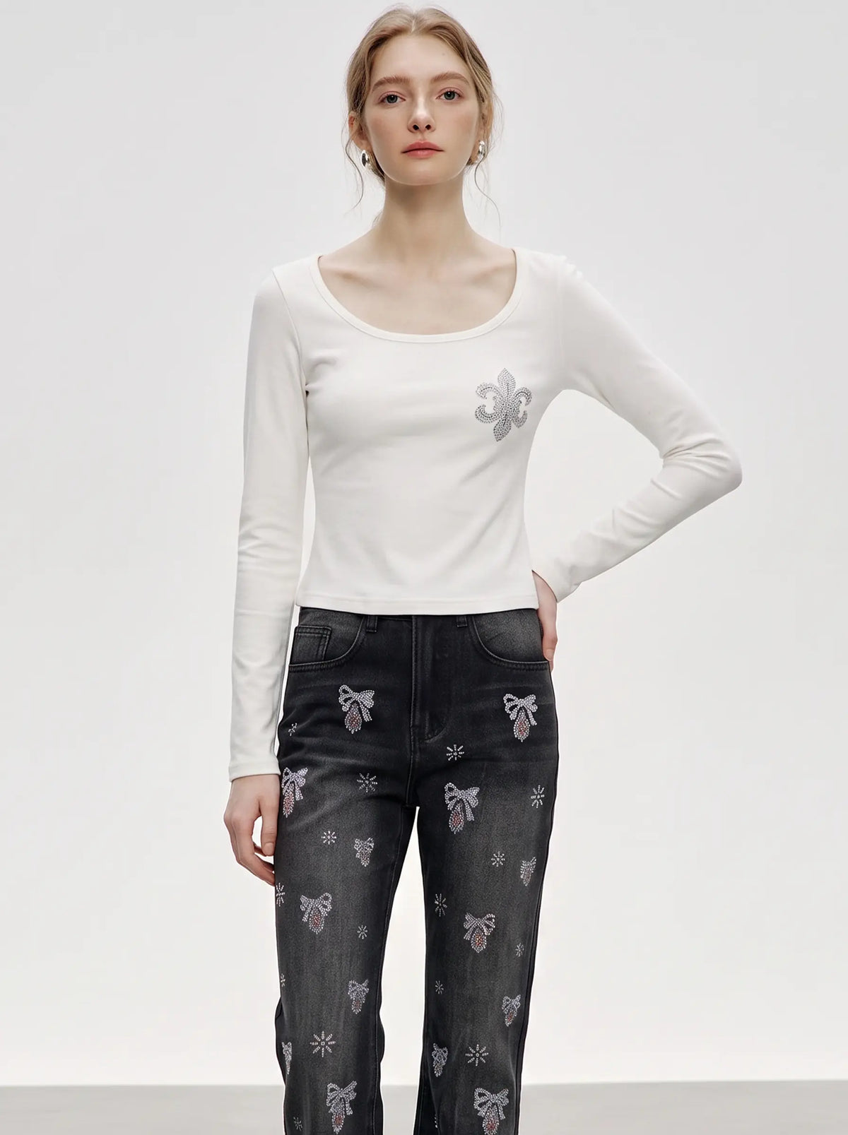 A model wearing a fitted white long-sleeve top with a silver fleur-de-lis embroidery on the left chest, paired with dark gray patterned jeans, against a plain light gray studio background. TCH Rhinestone Fleur-de-Lis Fitted Long Sleeve Top