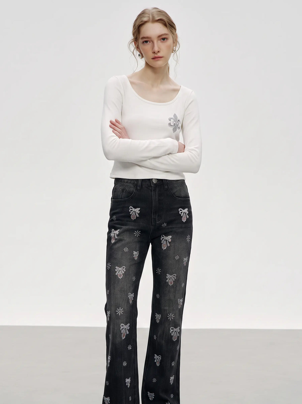 A model stands against a light gray studio background wearing a fitted white long-sleeve top with a small gray floral detail on the chest and dark gray wide-leg jeans covered in small gray floral and bow patterns. TCH Rhinestone Fleur-de-Lis Fitted Long Sleeve Top
