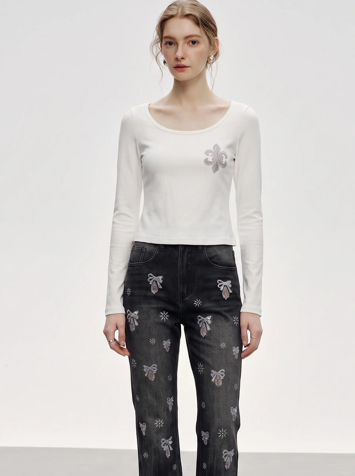A model stands against a light gray studio background wearing a fitted white long-sleeve top with a small gray bow embellishment on the chest and dark gray patterned jeans with bow motifs. TCH Rhinestone Fleur-de-Lis Fitted Long Sleeve Top