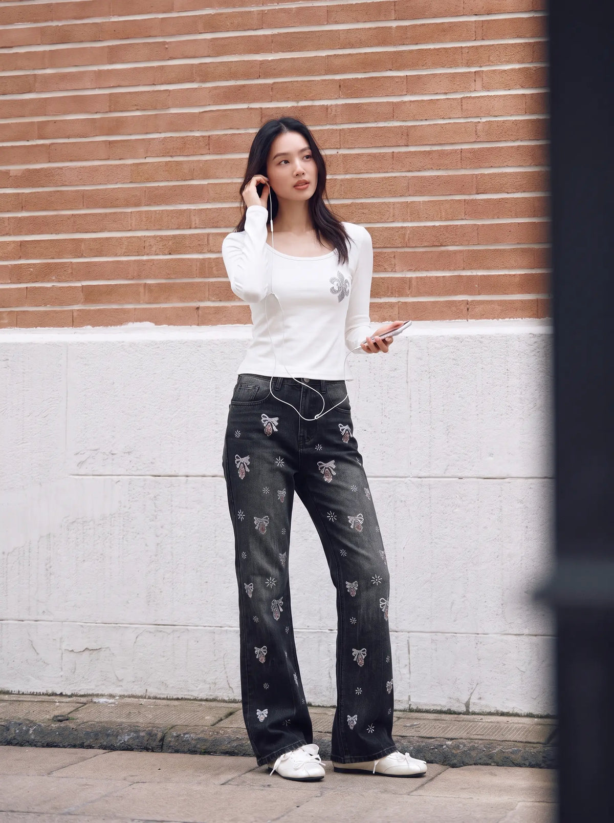 A woman stands outdoors against a brick and painted wall, wearing a fitted white long-sleeve top, dark patterned wide-leg pants, and white sneakers. TCH Rhinestone Fleur-de-Lis Fitted Long Sleeve Top