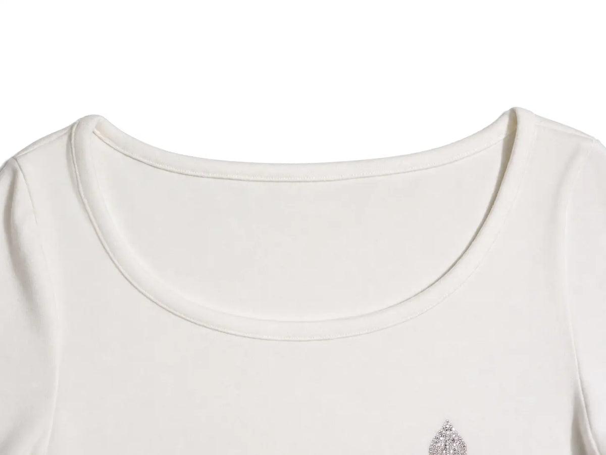 Close-up of a cream-colored scoop neck top with a small silver rhinestone embellishment on the lower right chest, set against a plain white background. TCH Rhinestone Fleur-de-Lis Fitted Long Sleeve Top