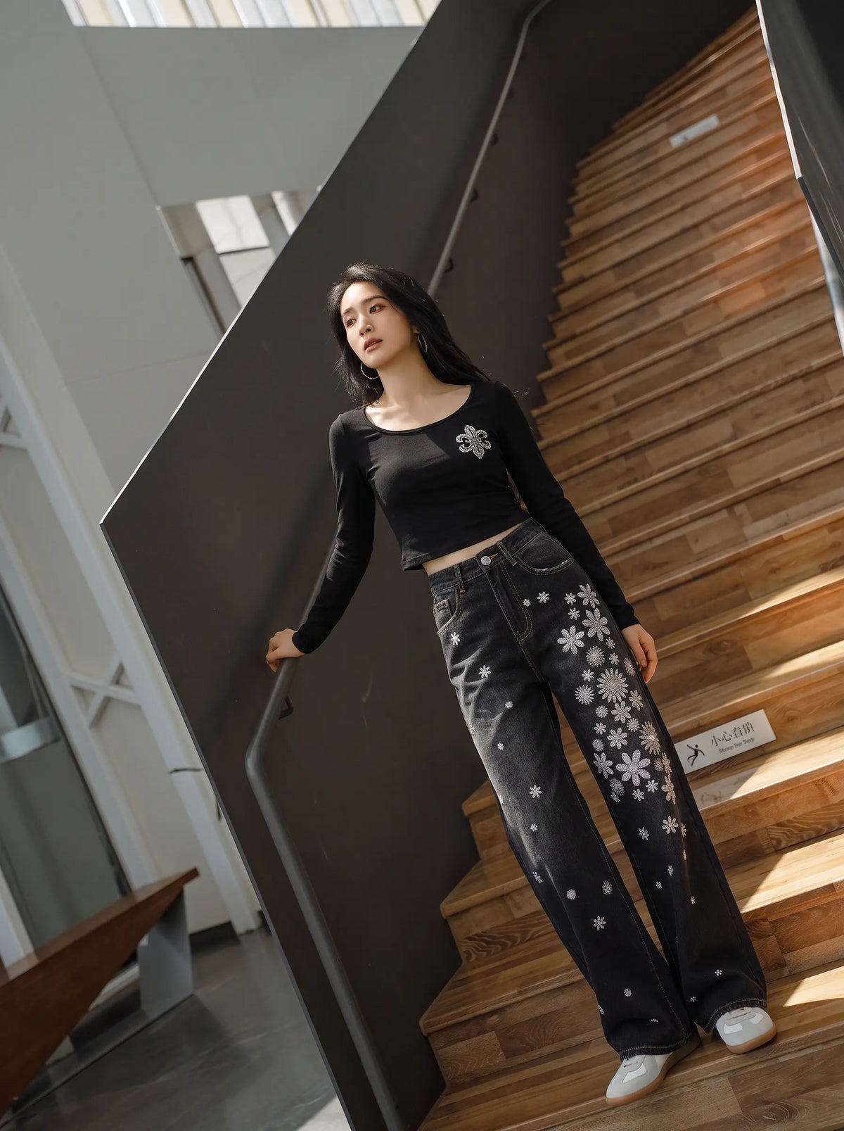 A woman stands on a wooden staircase wearing a black long-sleeve crop top with a silver graphic on the chest and black wide-leg jeans featuring white snowflake patterns down the left leg. TCH Rhinestone Fleur-de-Lis Fitted Long Sleeve Top