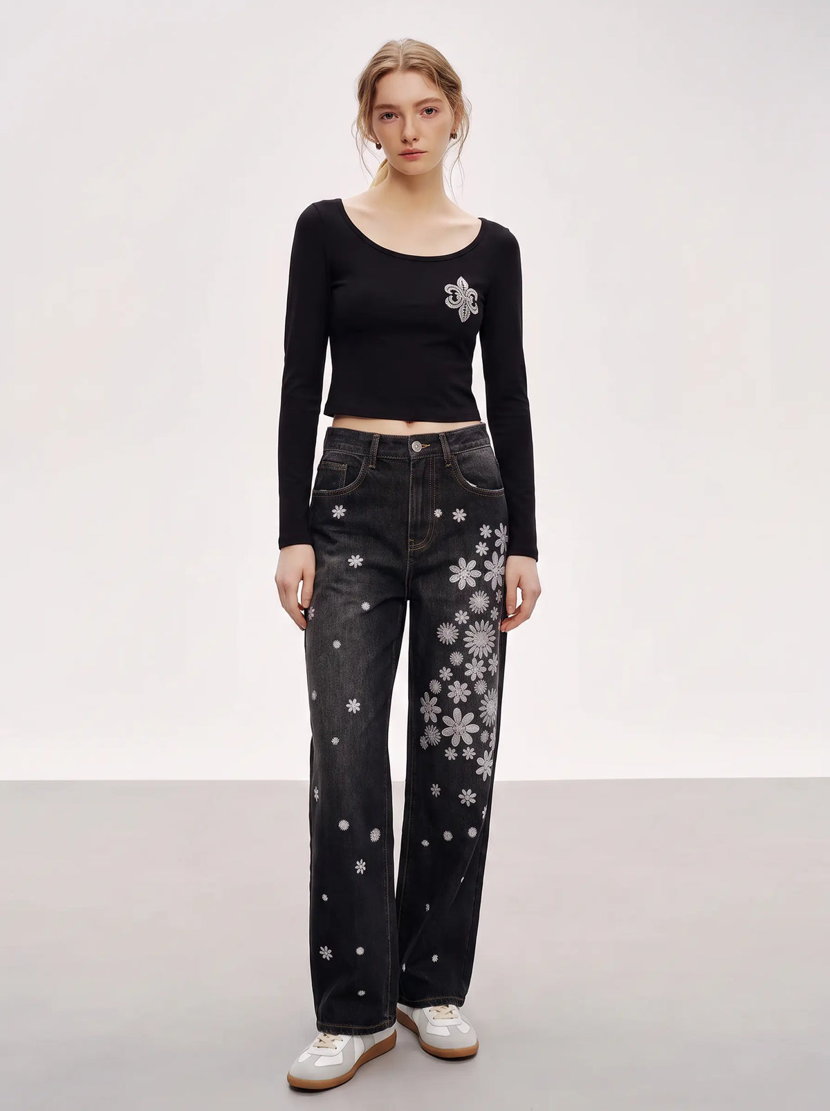 A model stands against a light gray studio background wearing a black long-sleeve crop top with a small white graphic on the chest, black jeans with white snowflake patterns, and white sneakers. TCH Rhinestone Fleur-de-Lis Fitted Long Sleeve Top